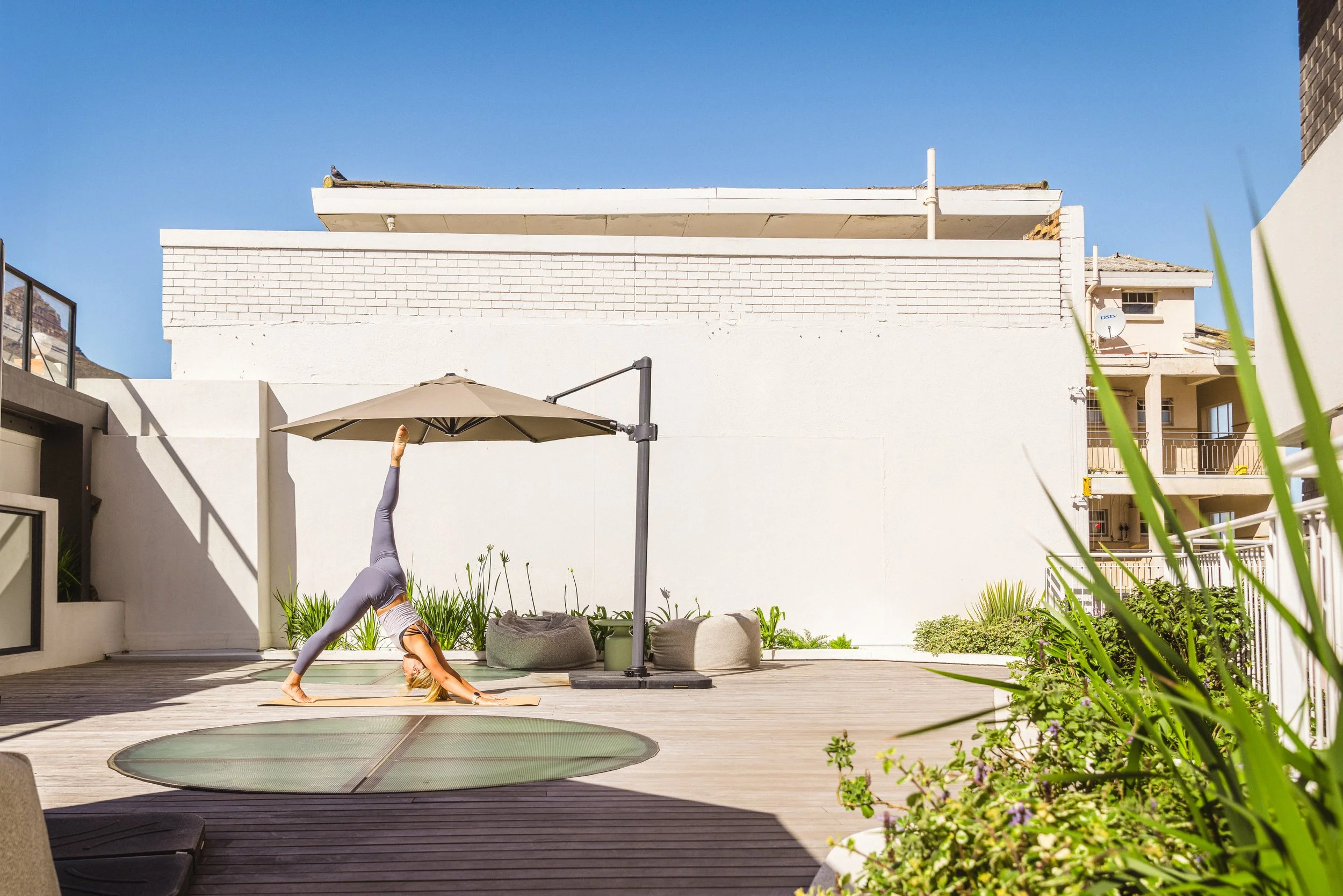 A woman practicing yoga on a rooftop patio in the sun, performing a downward dog pose with one leg raised, near an umbrella and some outdoor cushions, with a white wall and neighboring buildings in the background.
