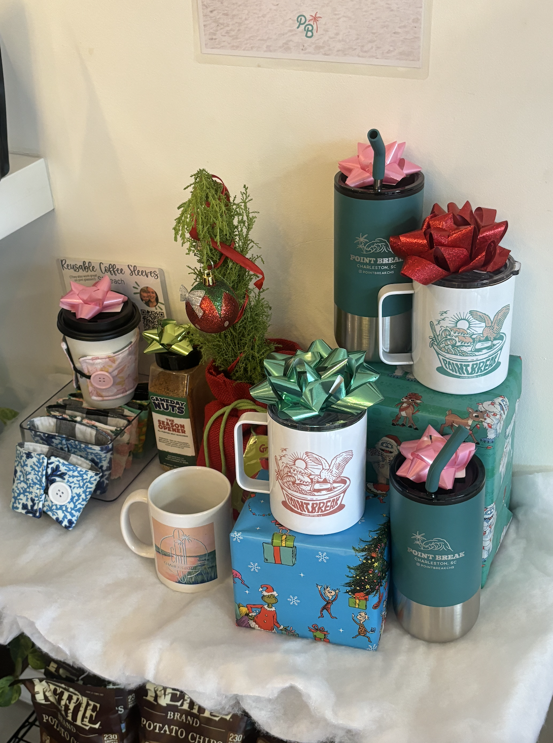A display of holiday gift items and travel mugs on a table, including small wrapped boxes, a miniature potted plant decorated with Christmas ornaments, reusable coffee sleeves, and ceramic mugs with holiday designs, all with colorful bows and wrapping paper.