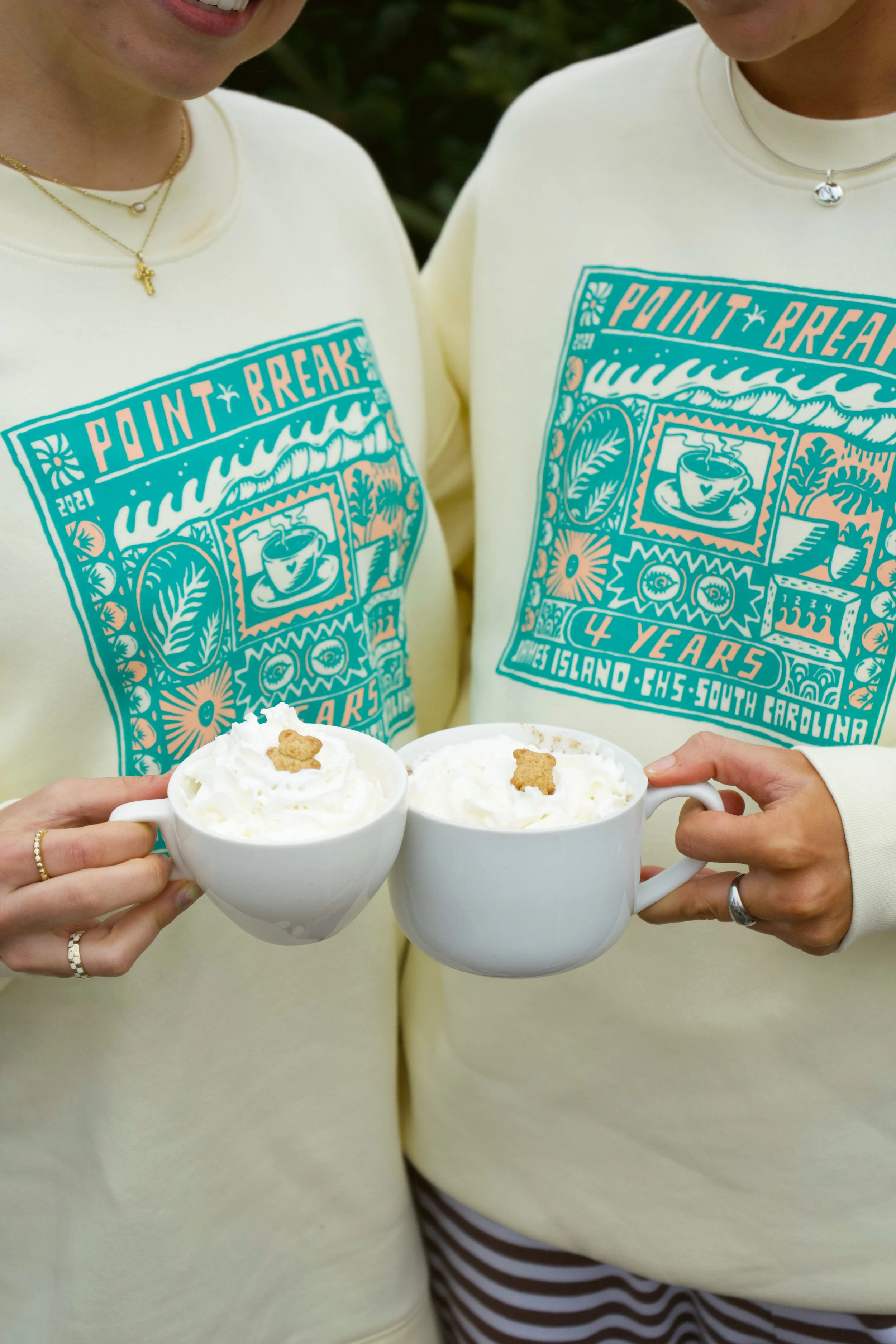 Two people holding white mugs with whipped cream and cookies, wearing matching yellow sweaters with a 'Point Break' 4-year anniversary graphic.