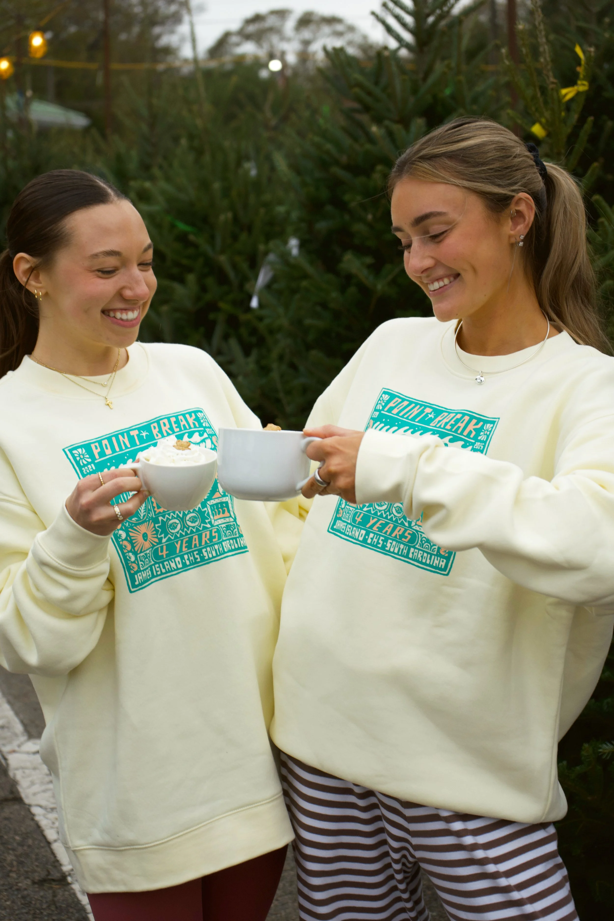 Two women wearing matching sweatshirt with a design for Point Break 4 Years James Island, Charleston, South Carolina, are smiling and clinking mugs and a bowl, enjoying a warm drink outdoors near Christmas trees.