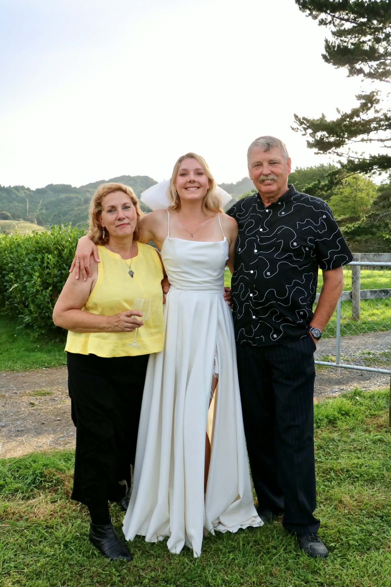 Three people standing outdoors on grass, posing for a photo during a celebration, with a young woman in a white dress in the center, flanked by an older woman in a yellow top and black pants on the left, and an older man in a black patterned shirt and black pants on the right, with trees and hills in the background and a slightly overcast sky.