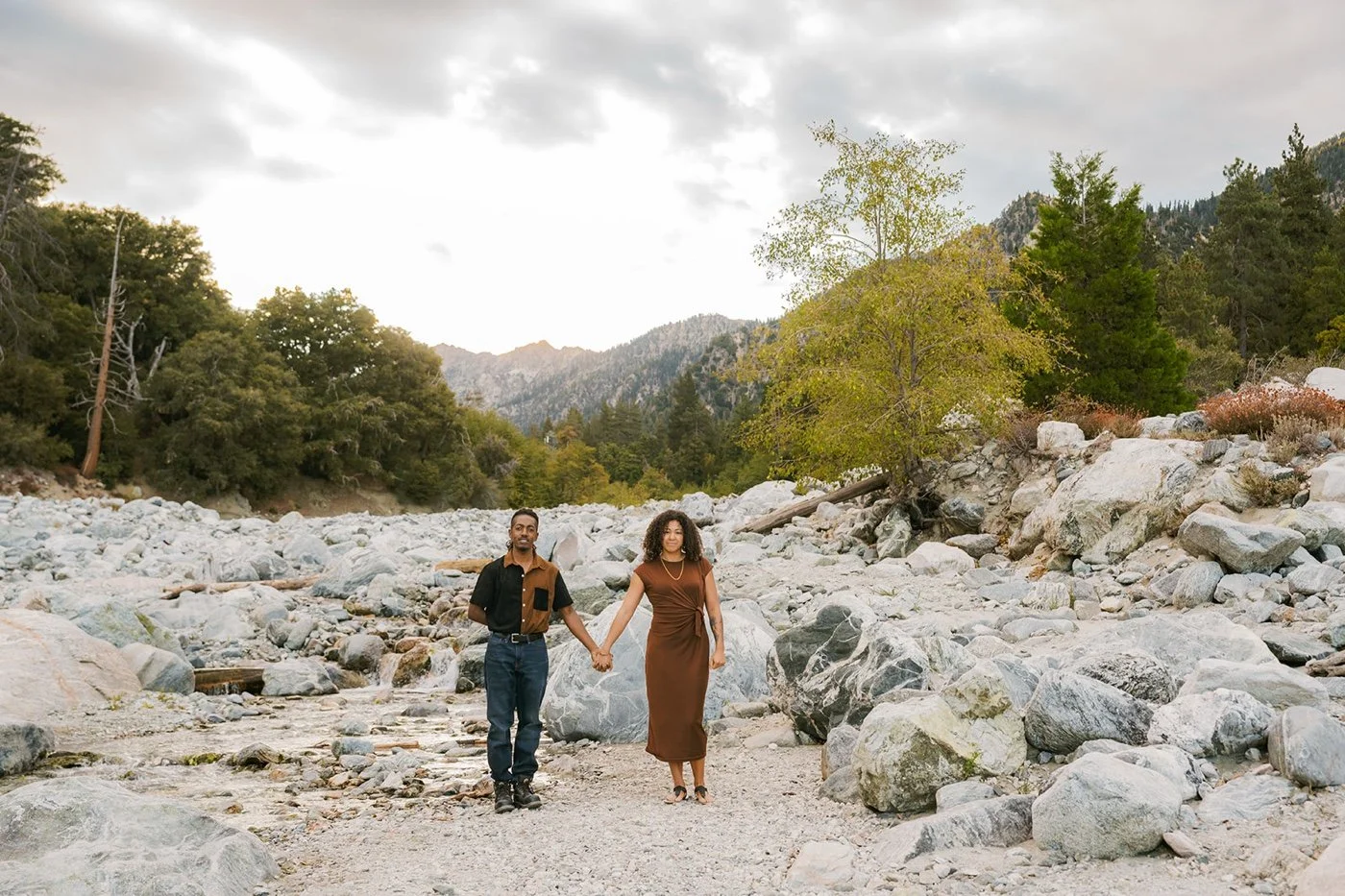a man and woman in earth toned clothes taking engagement photos by a river in forest falls