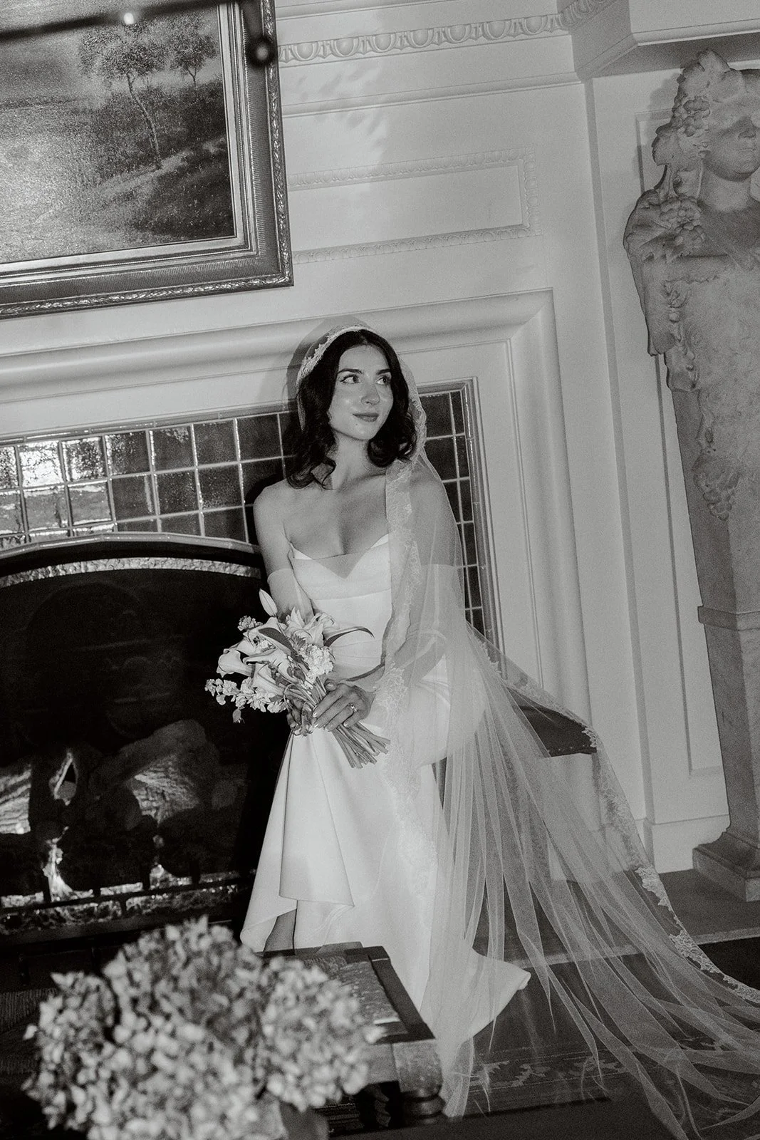 a black and white image of a bride wearing a veil holding her bouquet and sitting in a chair