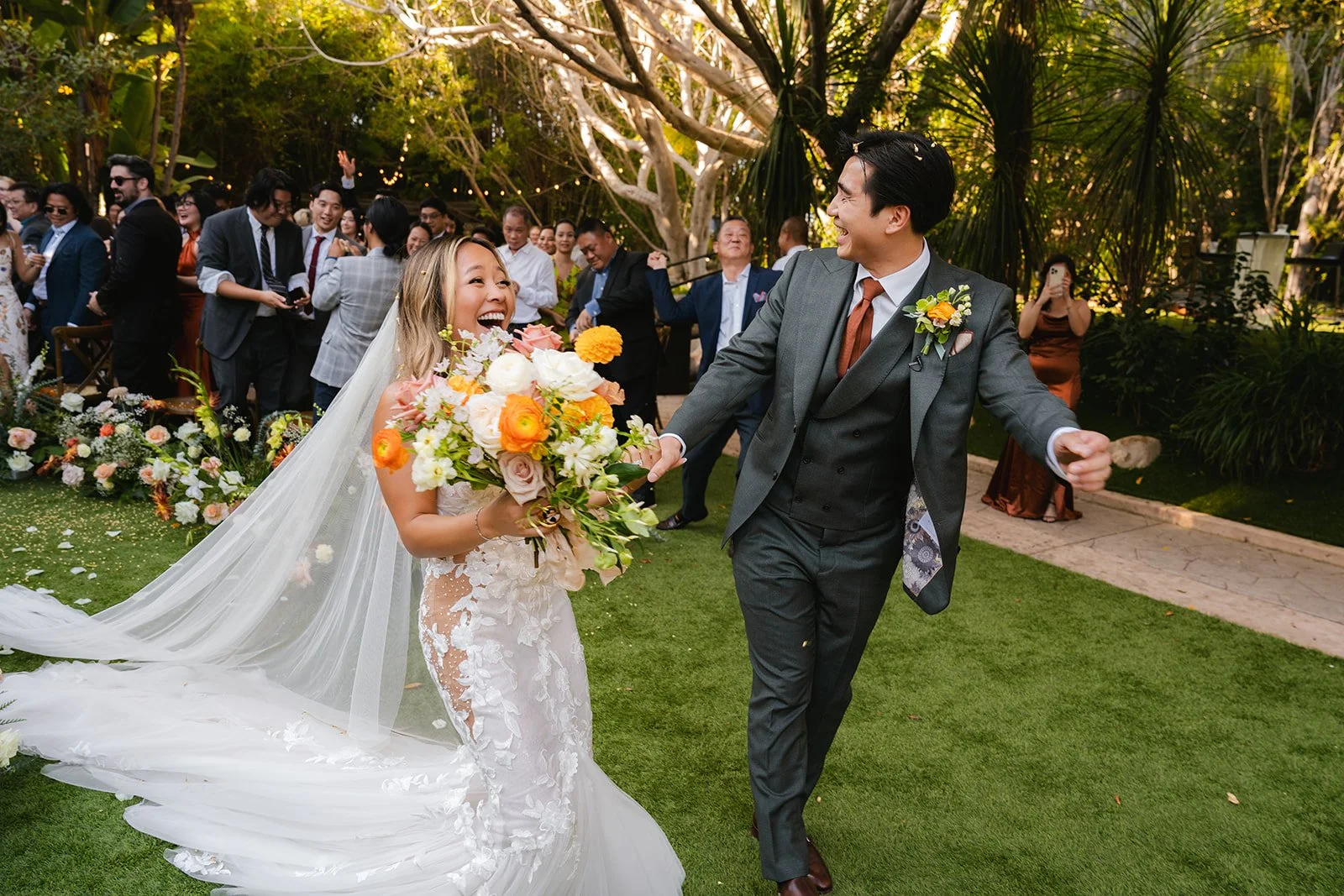 candid photo of a bride and groom walking down the aisle after getting married in a garden wedding, with the bride holding brightly colored flowers and the groom wearing a gray suit