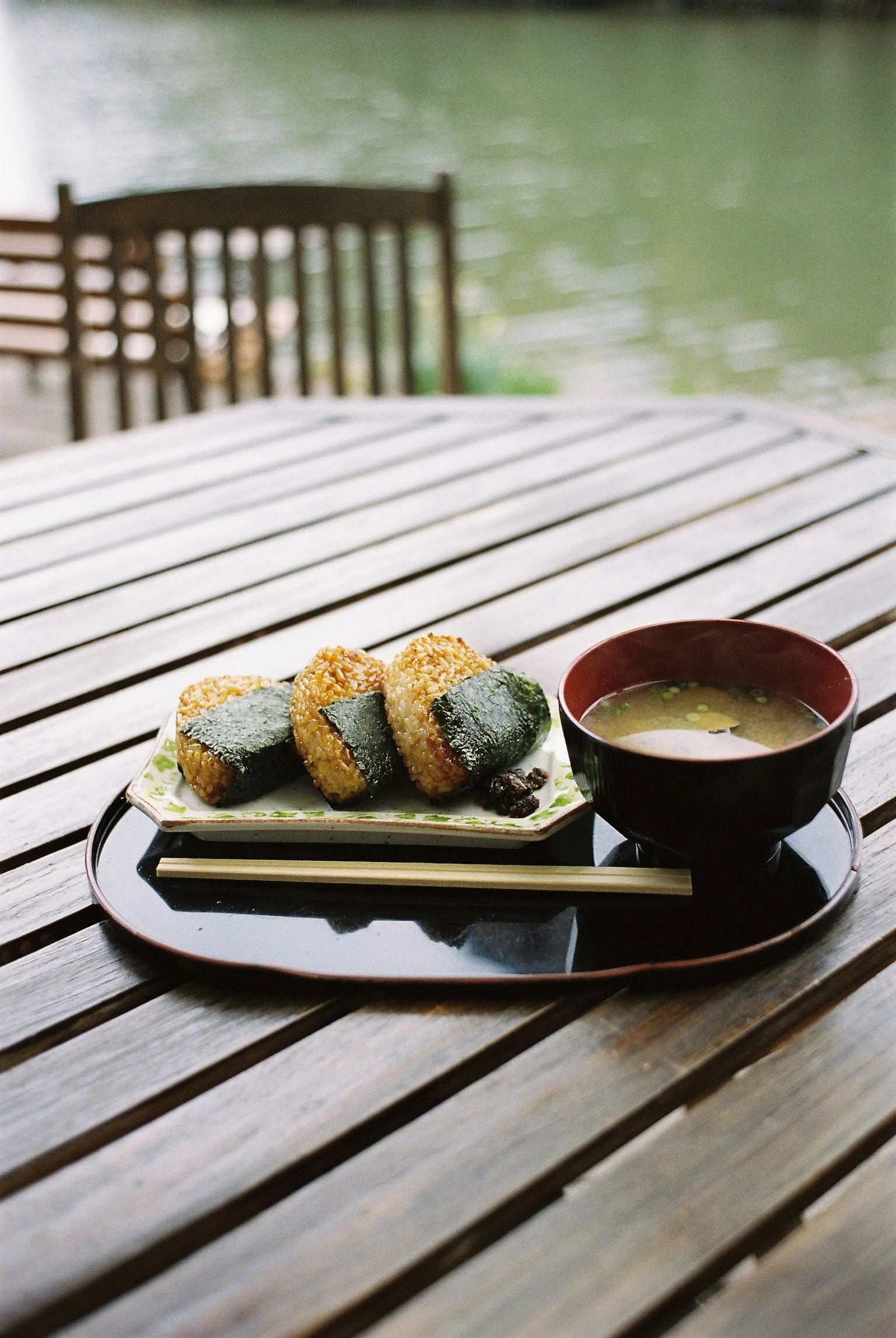 Bowl of noodle soup with meat and greens on a street food table, motorcycle with hanging bags and a vendor preparing food in the background.