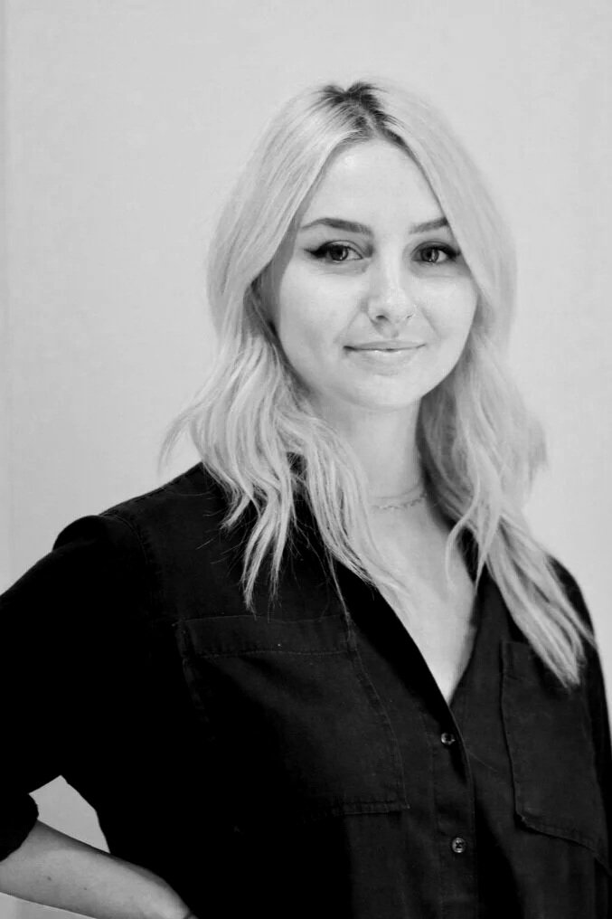 A woman with long wavy blonde hair, wearing a black shirt, smiling slightly, standing against a plain background.