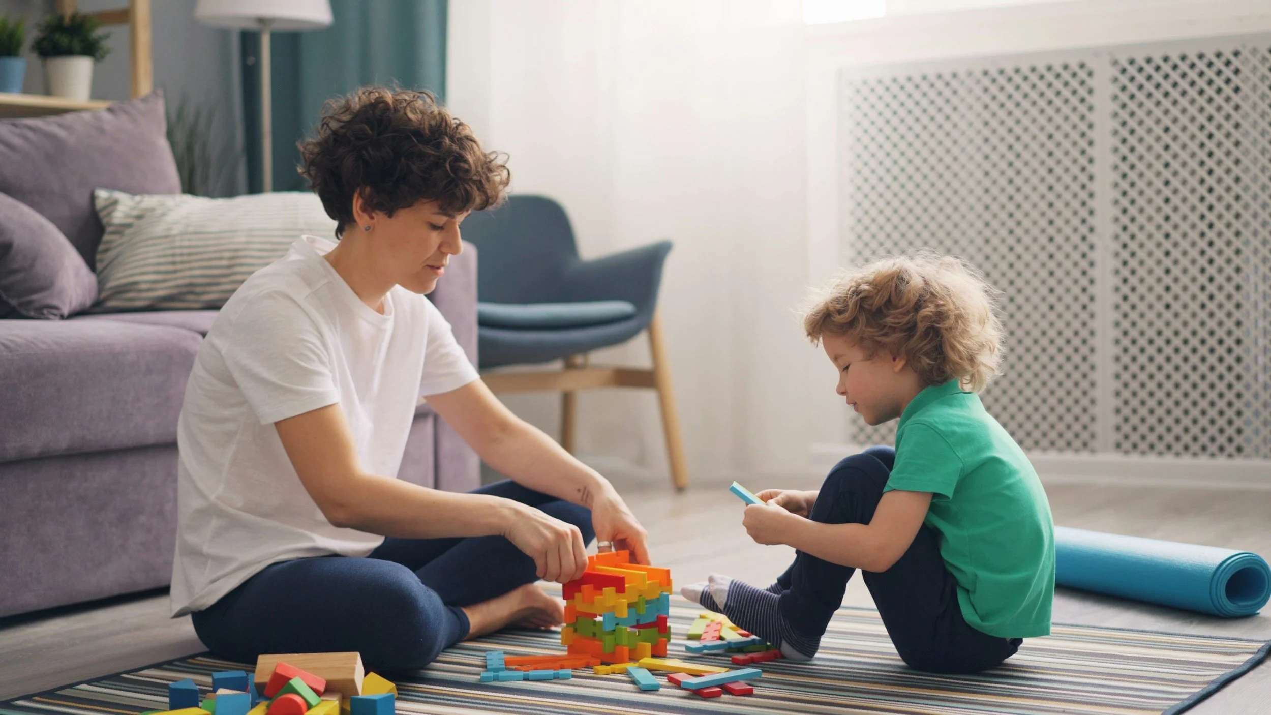 A woman and a young boy are sitting on a striped rug in a living room, playing with colorful building blocks. The woman is wearing a white t-shirt, and the boy is wearing a green shirt. There is a purple couch, a blue armchair, and a bookshelf in the background.
