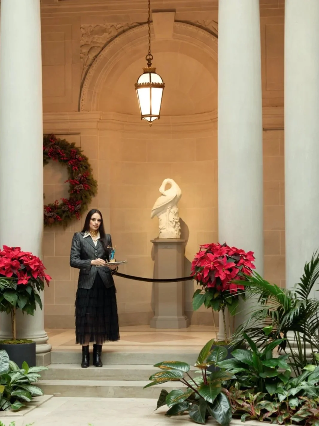 Woman in a dark suit stands with a tray and pens in front of a classical sculpture of a swan on a pedestal, inside a decorated building with red poinsettia plants, a wreath, columns, and a ceiling lantern.