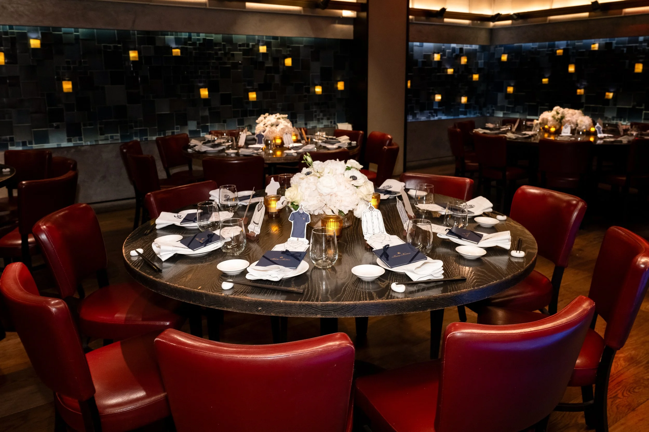 Elegant restaurant with round tables decorated with white floral centerpieces, surrounded by red leather chairs, and set with glasses, napkins, and small candles. Dark decorative wall in the background.