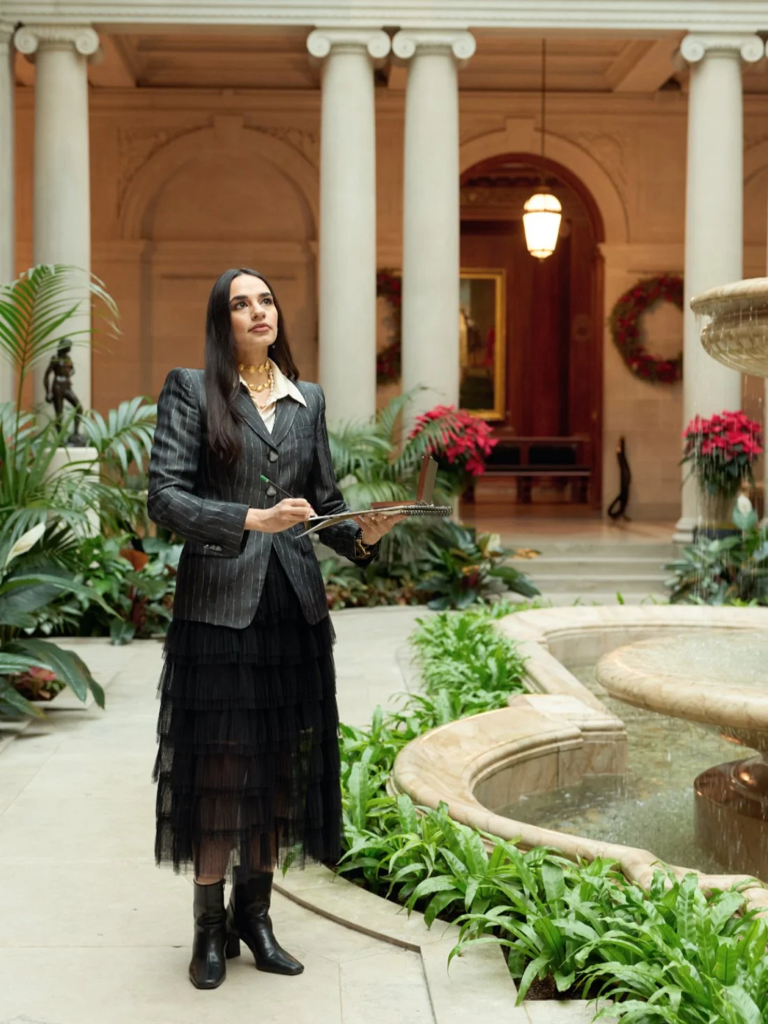 Woman in a pinstripe blazer and layered black skirt holding a notebook and pen standing in an elegant indoor garden with lush green plants, a fountain, and Christmas wreaths.