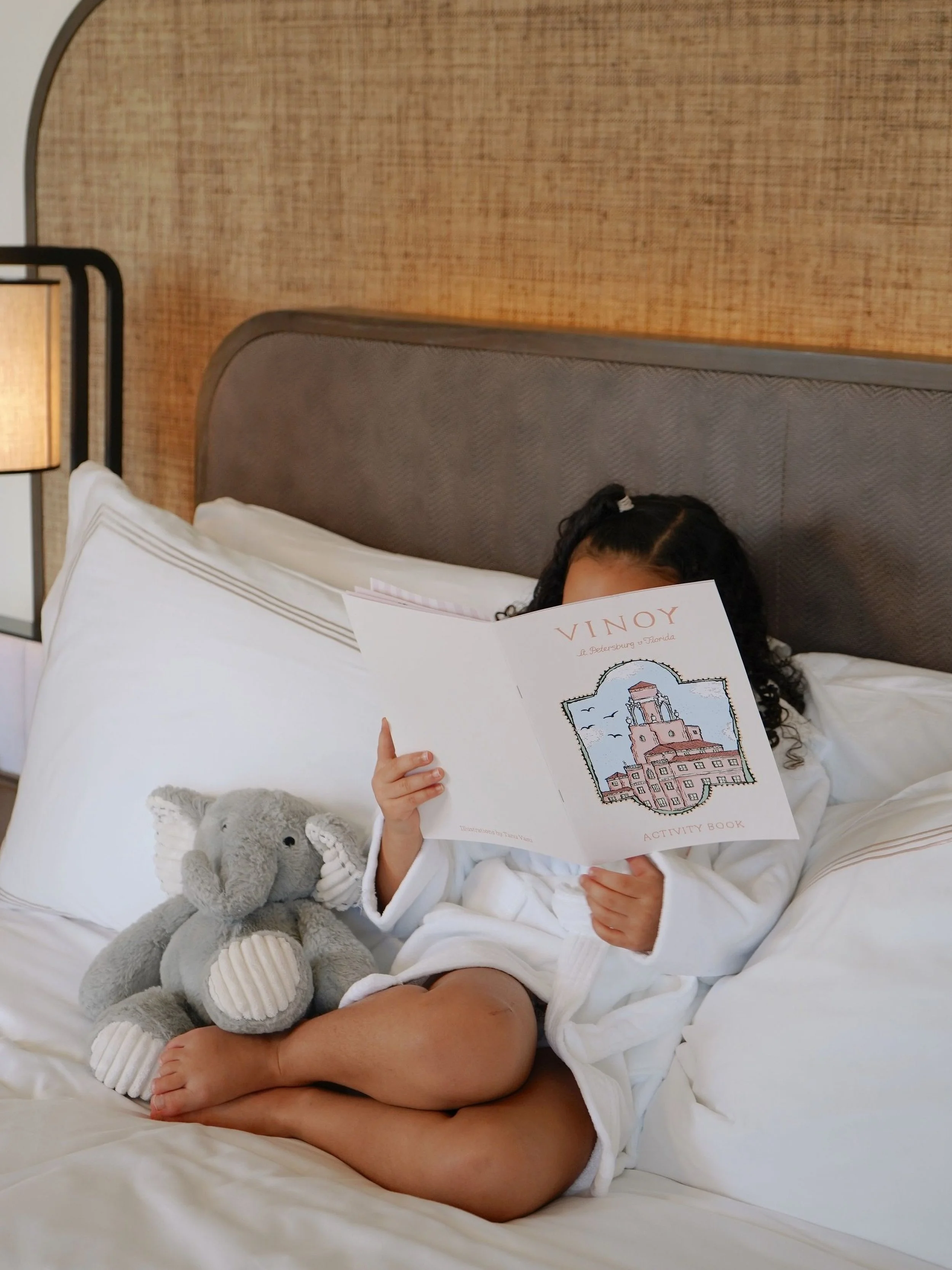 A child with curly hair is lying on a bed, holding a booklet titled 'VINOY' and an image of a building on the cover. The child also has a stuffed elephant toy beside them.