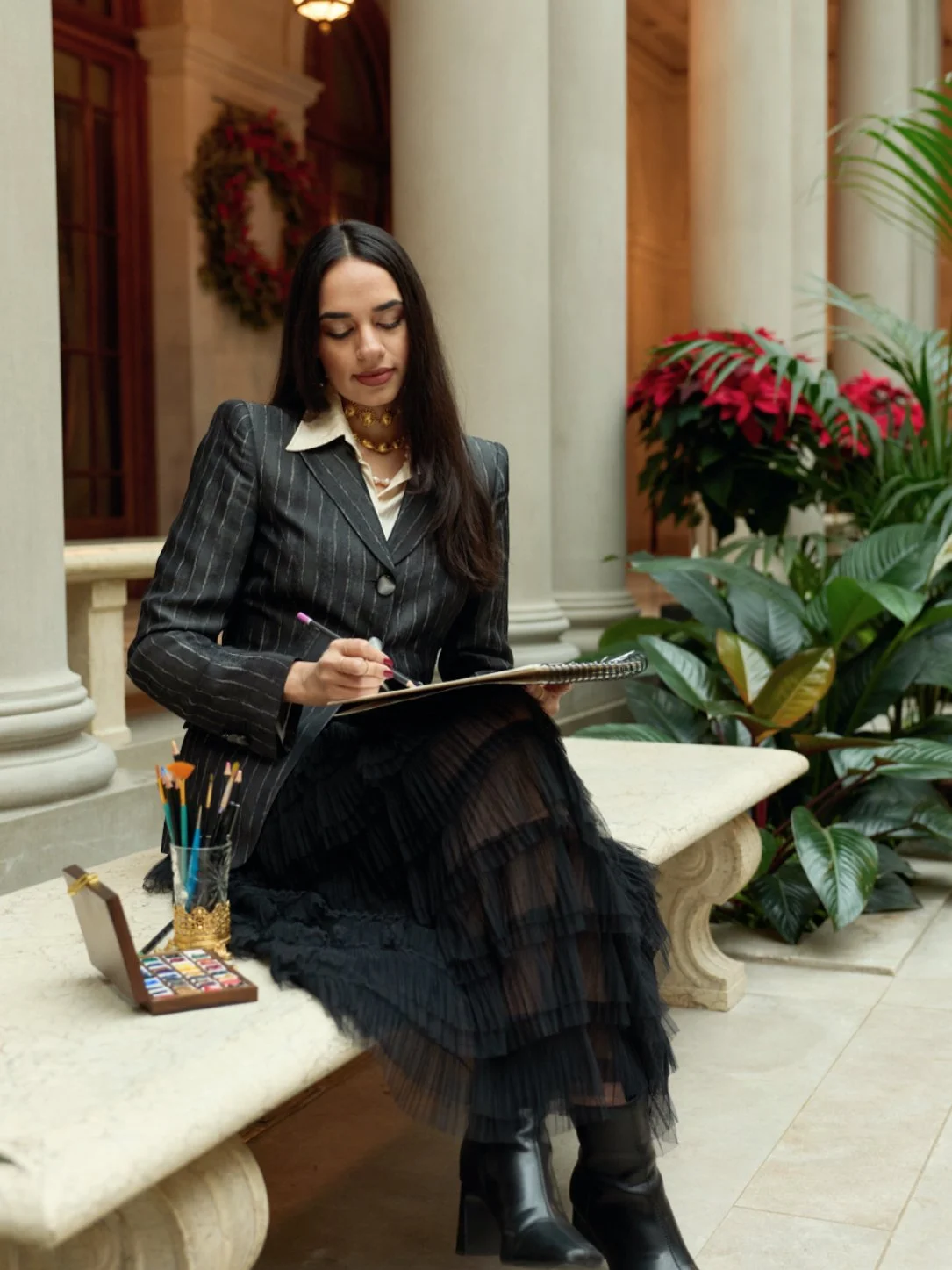 A woman in a pinstripe blazer and black skirt sitting on a marble bench, writing in a notebook. There are plants and a floral wreath in the background.