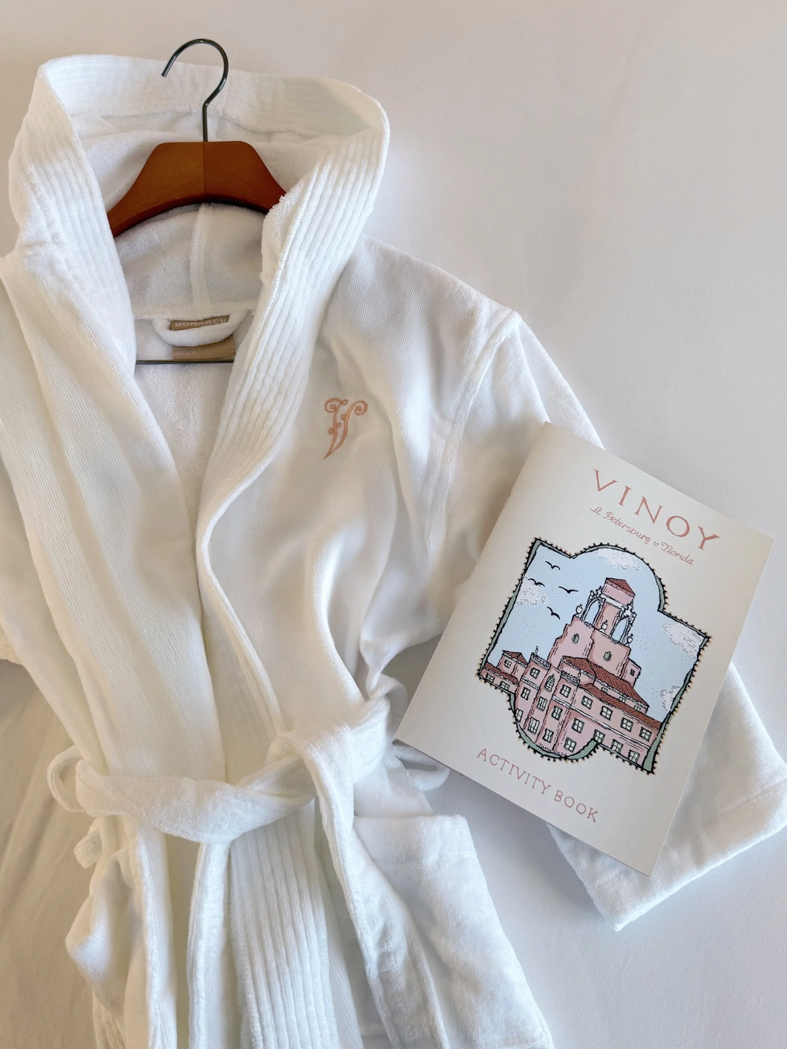 White bathrobe hanging on a wooden hanger with a small embroidered logo, next to a booklet with a drawing of a historic building and the words "VINOY ... ACTIVITY BOOK."