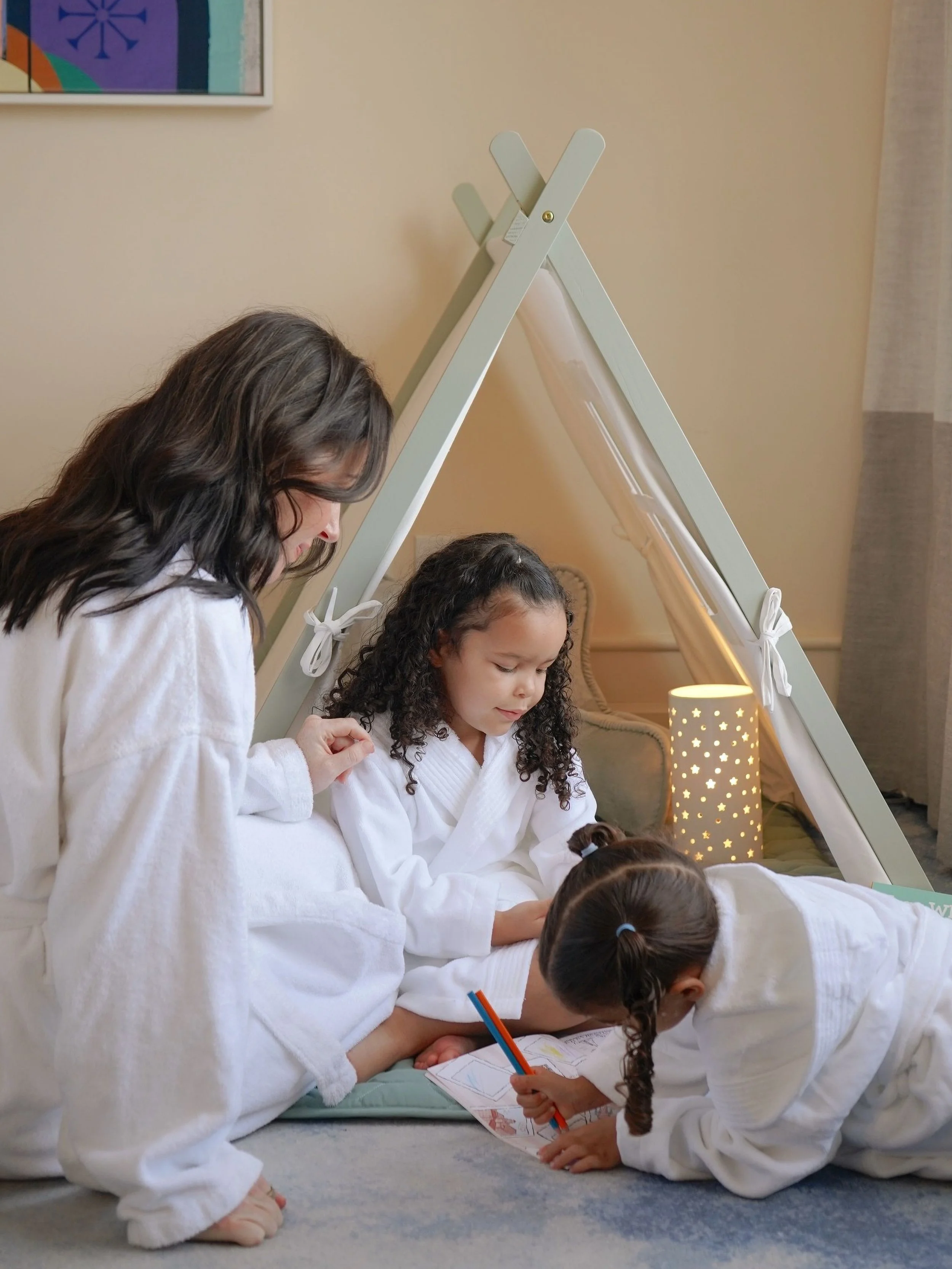 Children dressed in white robes, participating in a group activity, with some sitting and some lying on the floor, inside a room with beige walls and a colorful artwork on the wall.