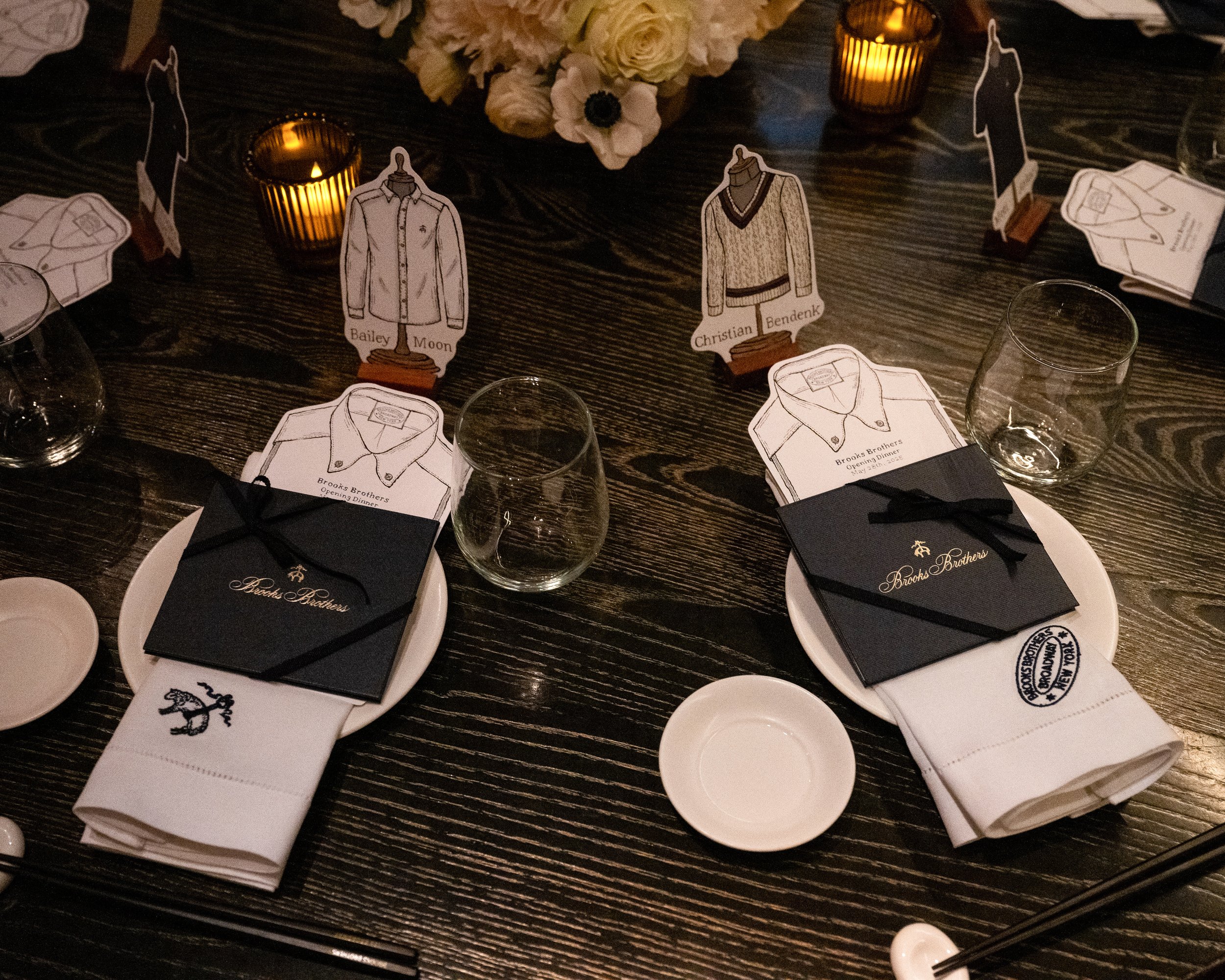 Table with decorative paper clothing cutouts labeled 'Bailey Moon' and 'Christian Bendenk,' surrounded by plates, glasses, candle holders, and floral centerpiece, indicating a formal dinner or event setting.