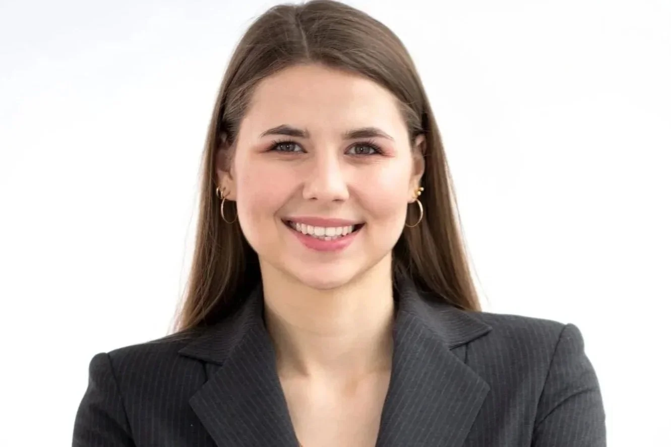 A woman with long brown hair, wearing a black blazer, and gold hoop earrings, smiling against a plain white background.