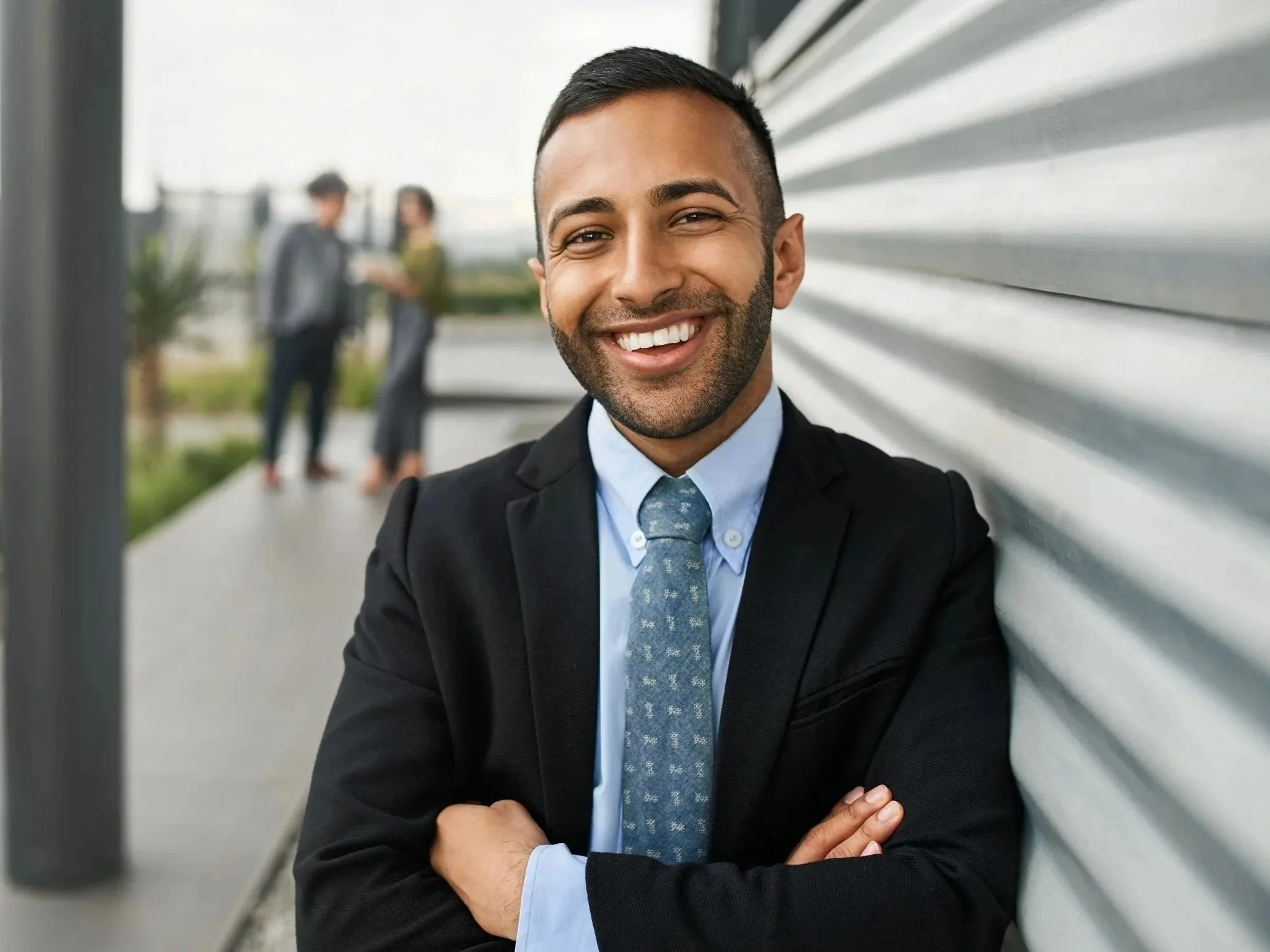A smiling man in a business suit and tie taking a selfie outdoors. Two people are blurred in the background, standing on a terrace or balcony with a metal and horizontal siding wall.