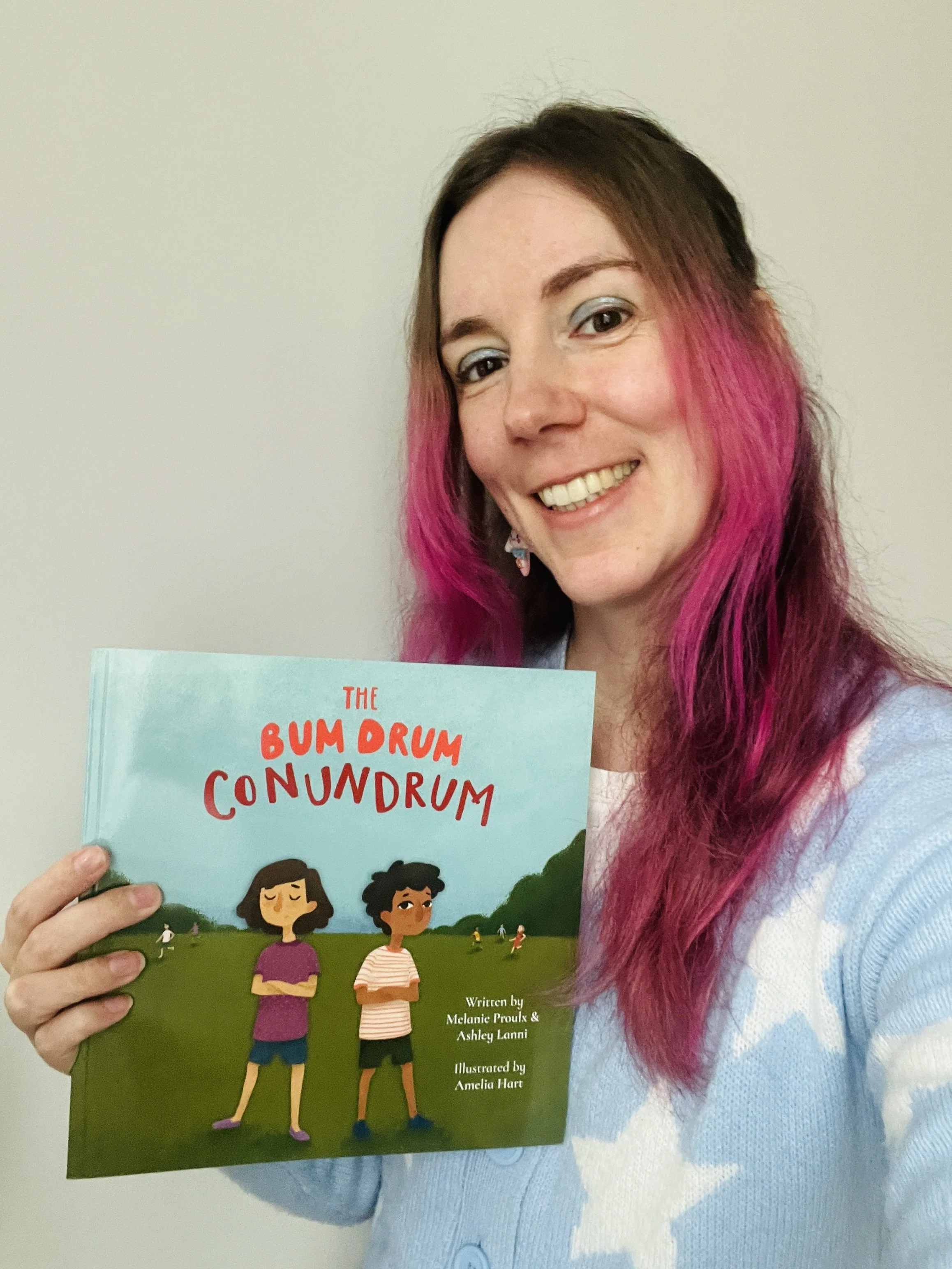 A woman with bright pink hair wearing a blue sweater with white stars holds up a picture book, The Bum Drum Conundrum.