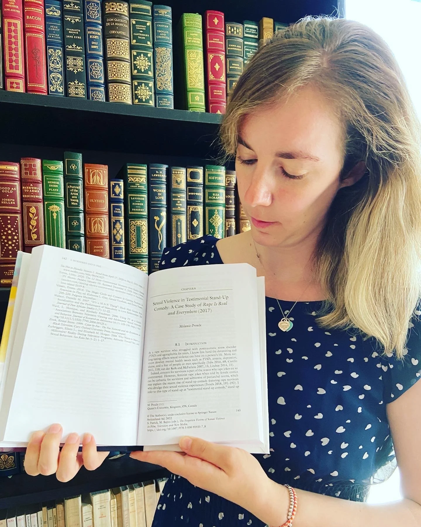 A woman with blond hair wearing a black dress with small white hearts looks down at a book she is holding. The book is open to the book chapter "Sexual Violence in Testimonial Stand-Up Comedy"