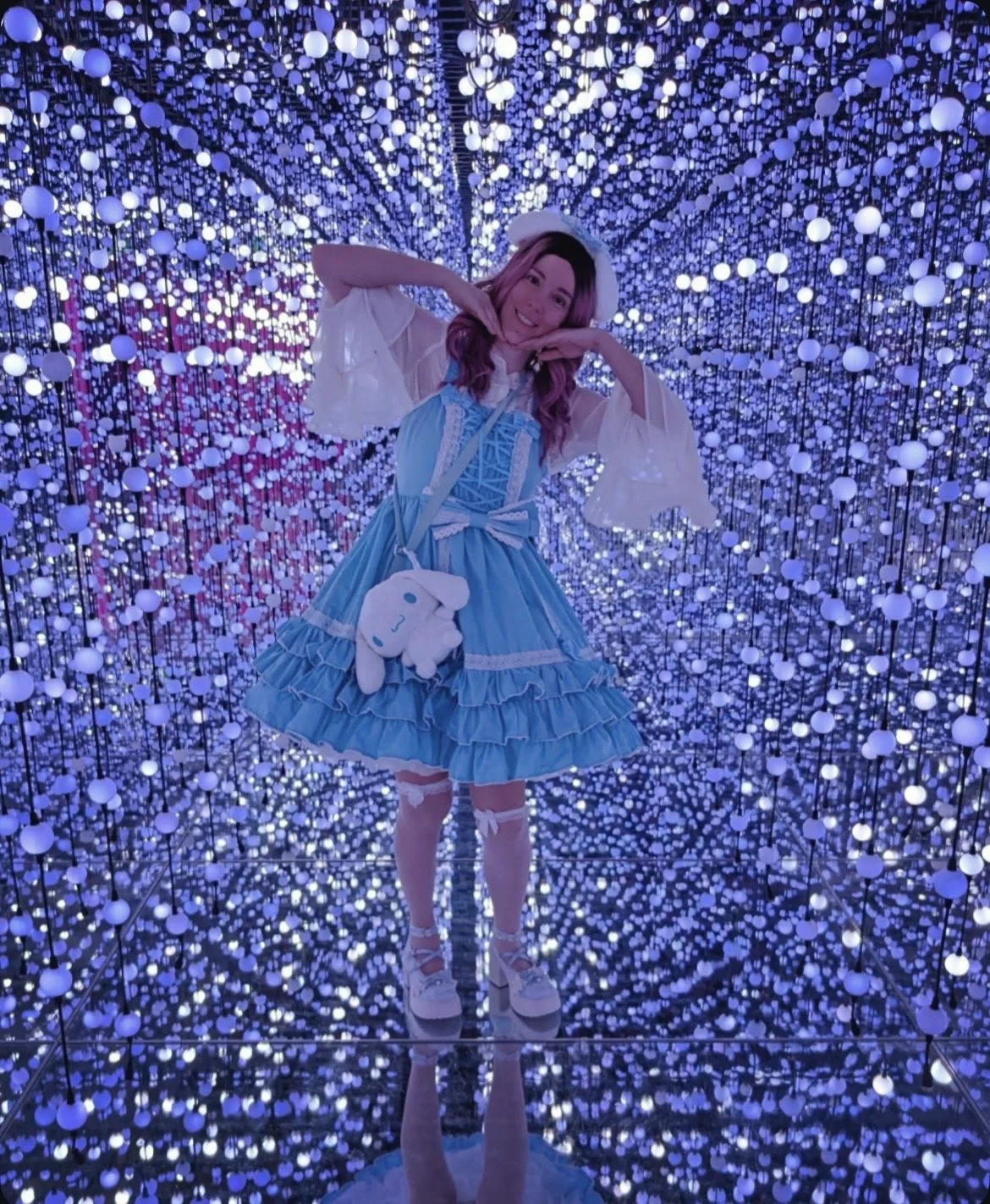 A woman picture book author wearing a lacy blue skirt and matching heels stands in a corridor full of bright lights. She is tilts her head to the side and holds her hands under her chin in a cute pose. She also has a cinnamoroll purse.