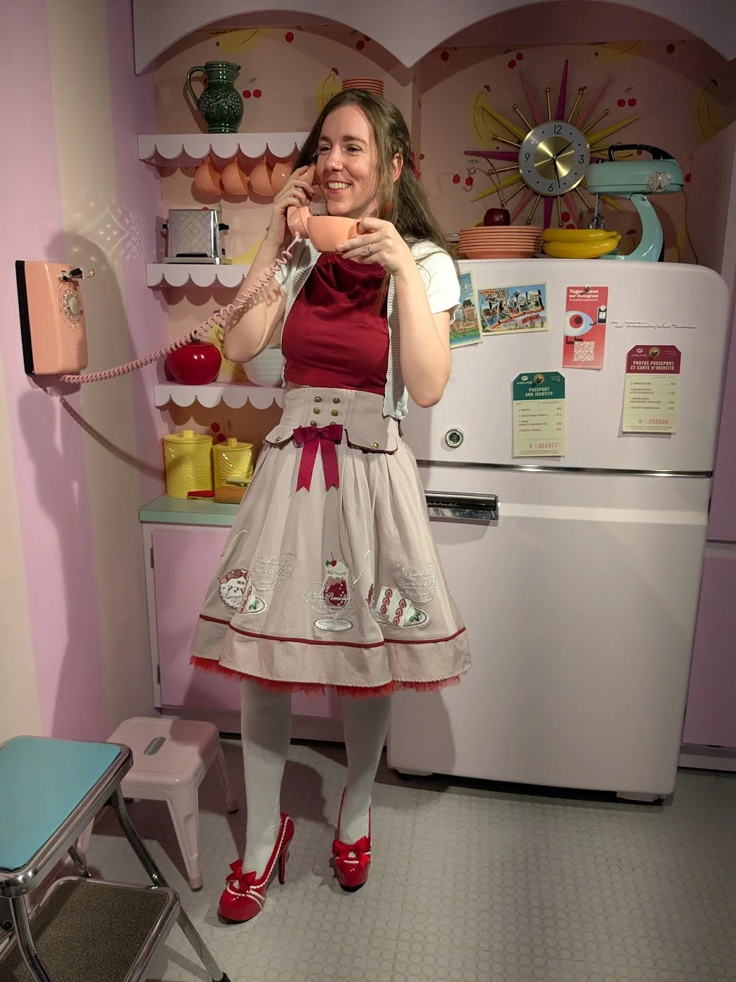 A woman picture book author wearing a puffy pink skirts with desserts embroidered on the bottom stands in a retro pink kitchen. She holds a coral pink phone to hear ear with one hand and a matching tea cup in the other.