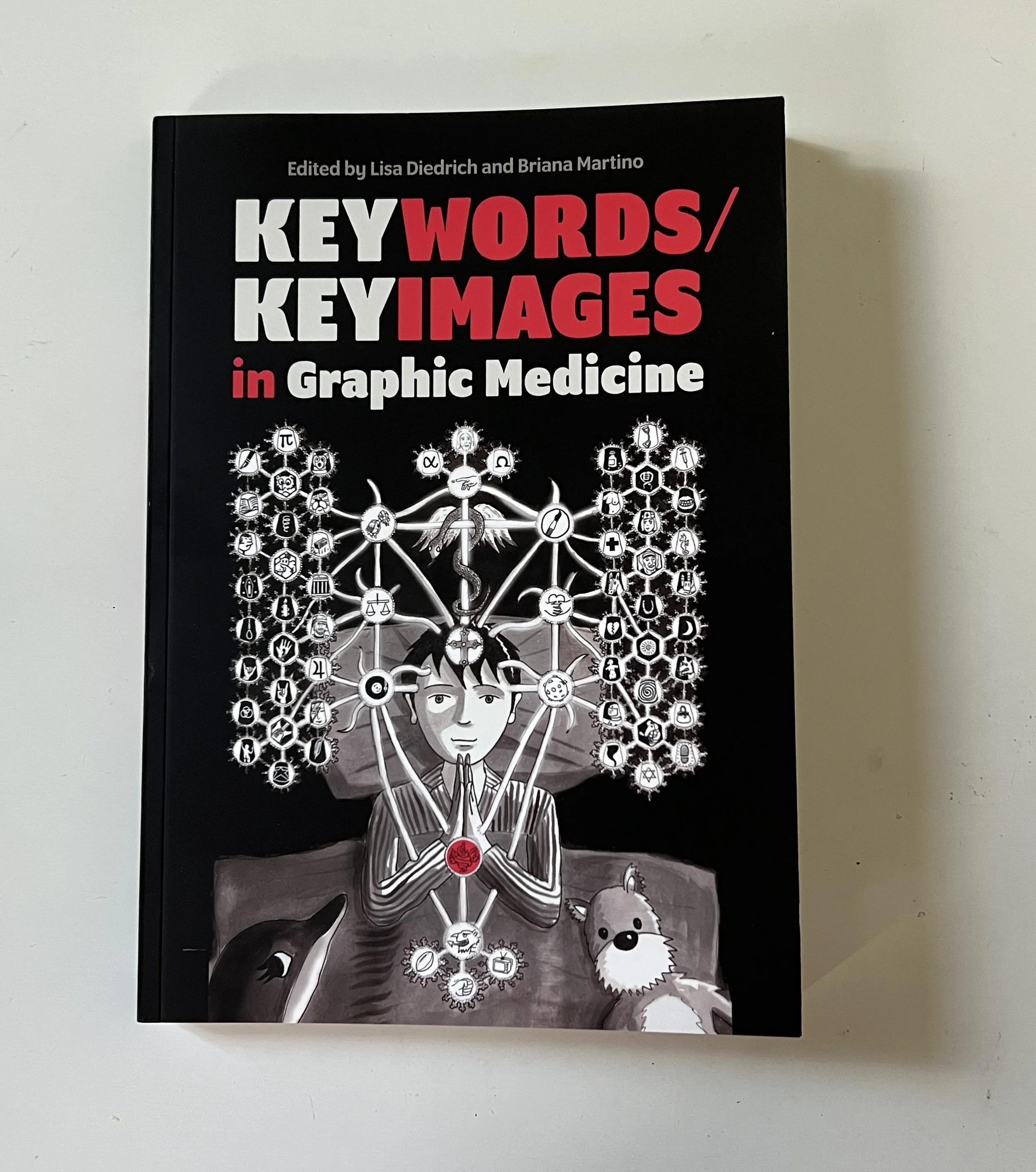 The cover of Keywords and Keyimages in Graphic Medicine. A boy lies in bed with mysterious symbols surrounding him as he bows his hands above his heart.