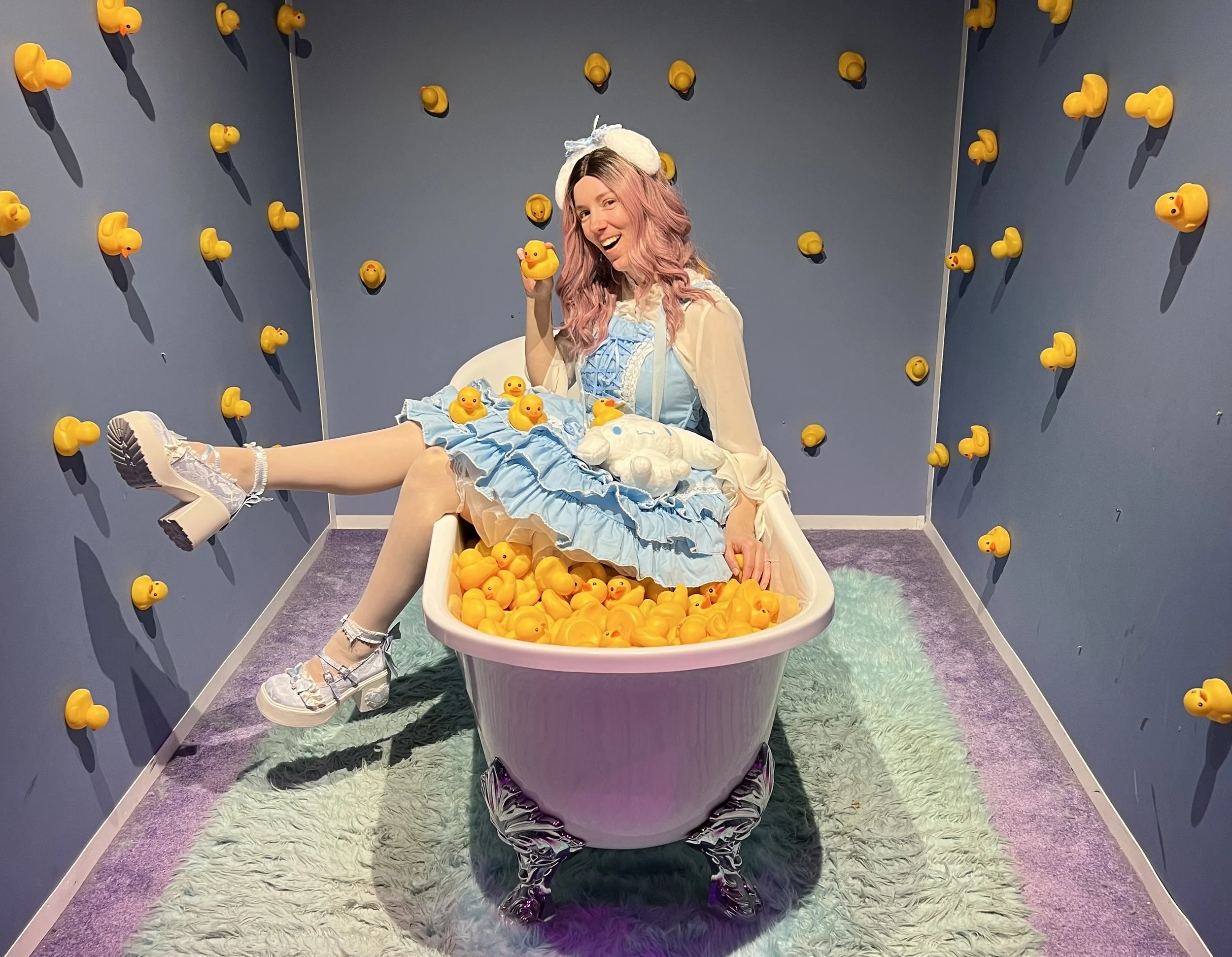 A woman picture book author wears a blue, lacy, puffy dress while sitting in a bathtub full of plastic yellow ducks. She has pink hair and is holding up a duck in one hand.