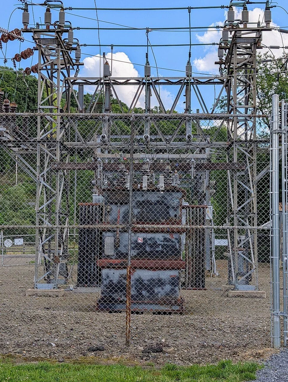 Electrical transformer and power lines behind a chain-link fence at an outdoor substation.