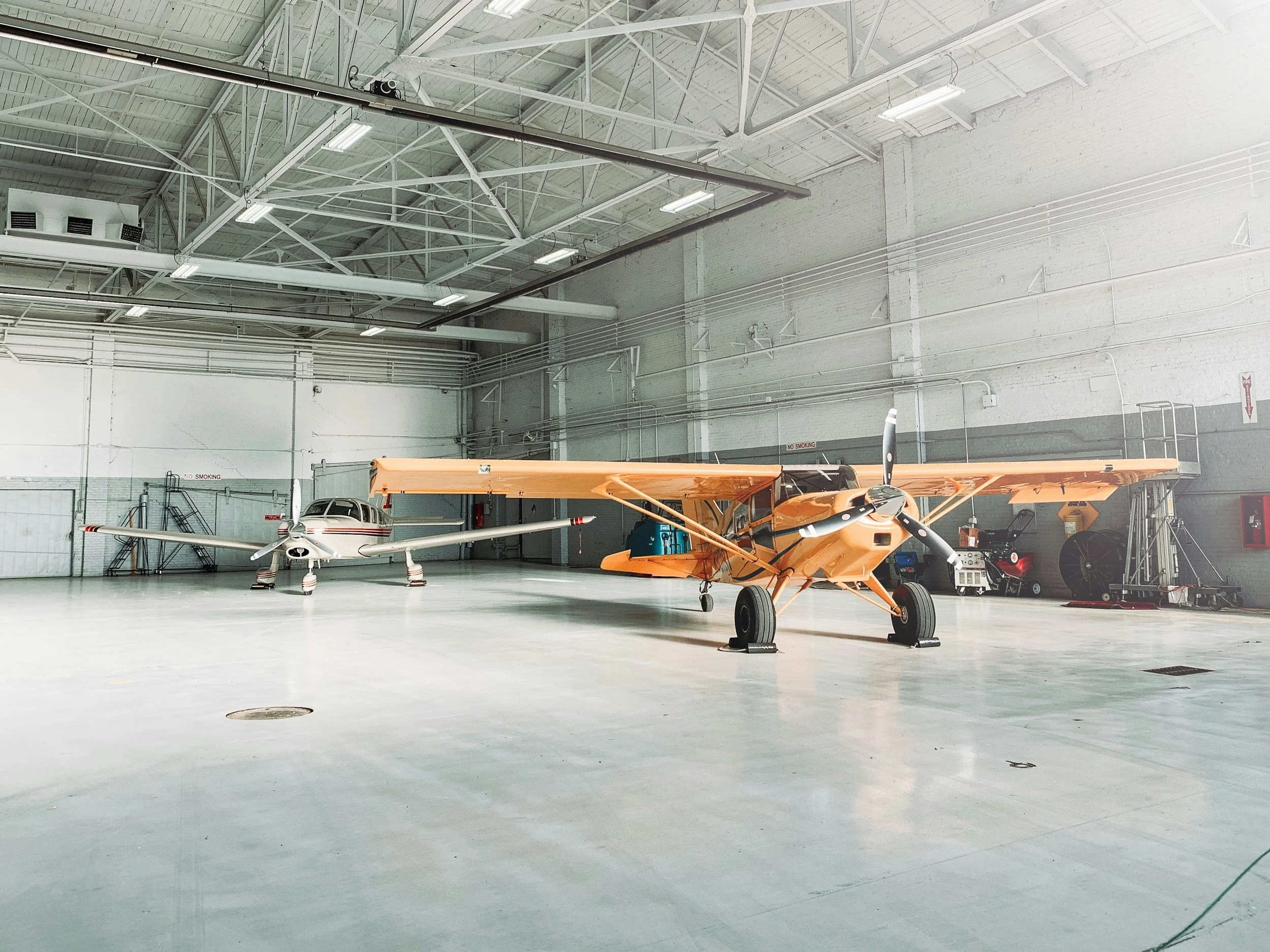 Two small airplanes inside a large hangar with white walls and high ceiling, one orange and one white. The hangar has a concrete floor and various equipment along the walls.