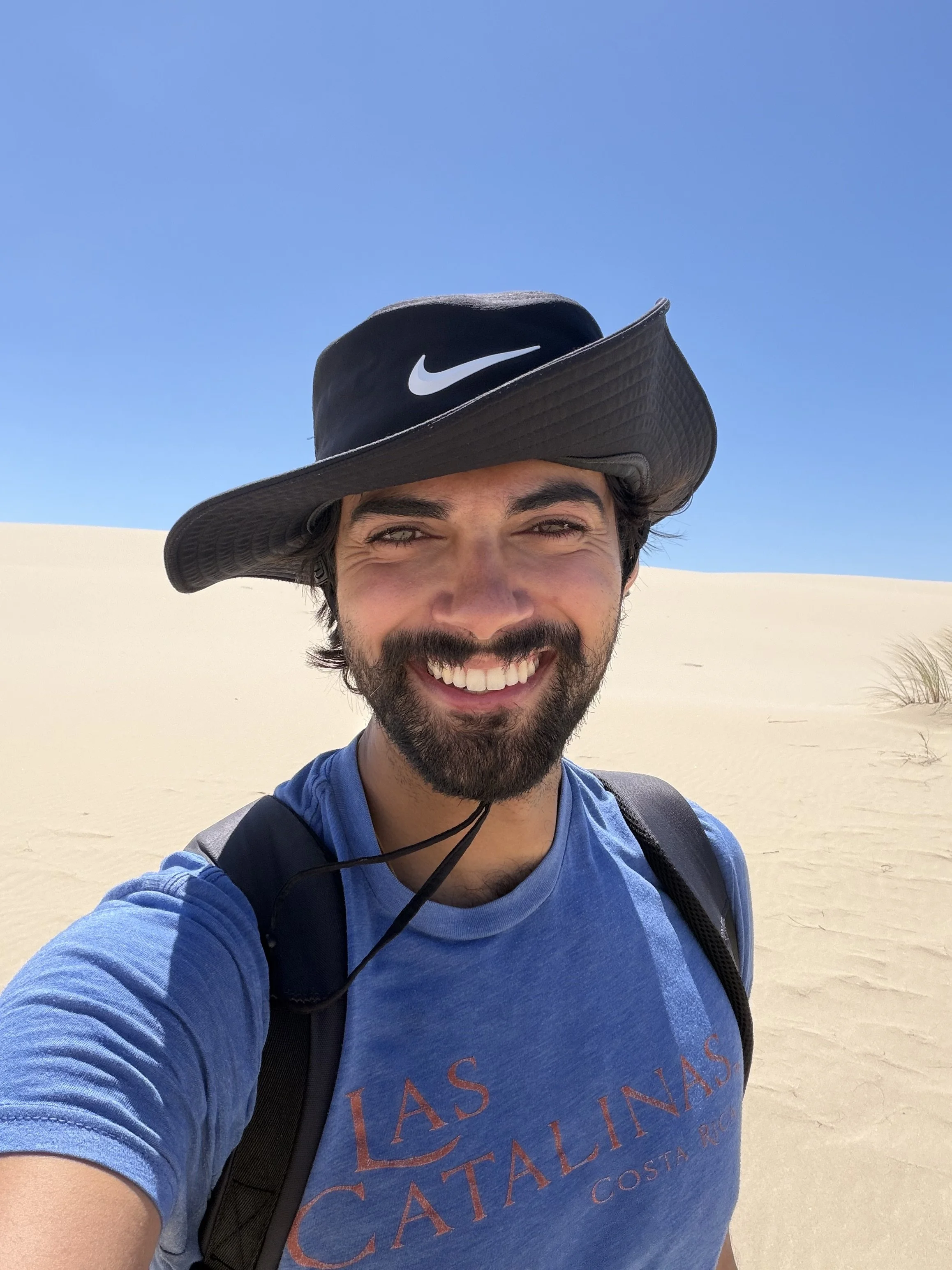 Nathan wearing a wide-brimmed Nike hat, blue T-shirt, and carrying a backpack, standing in a desert with white sand dunes and a clear blue sky.