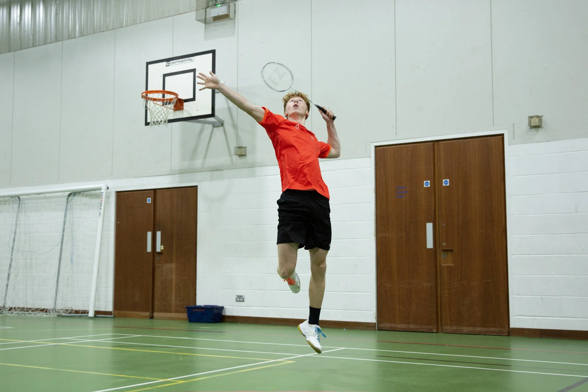 A young man playing badminton indoors, hitting the shuttlecock towards the net, with a basketball hoop in the background.