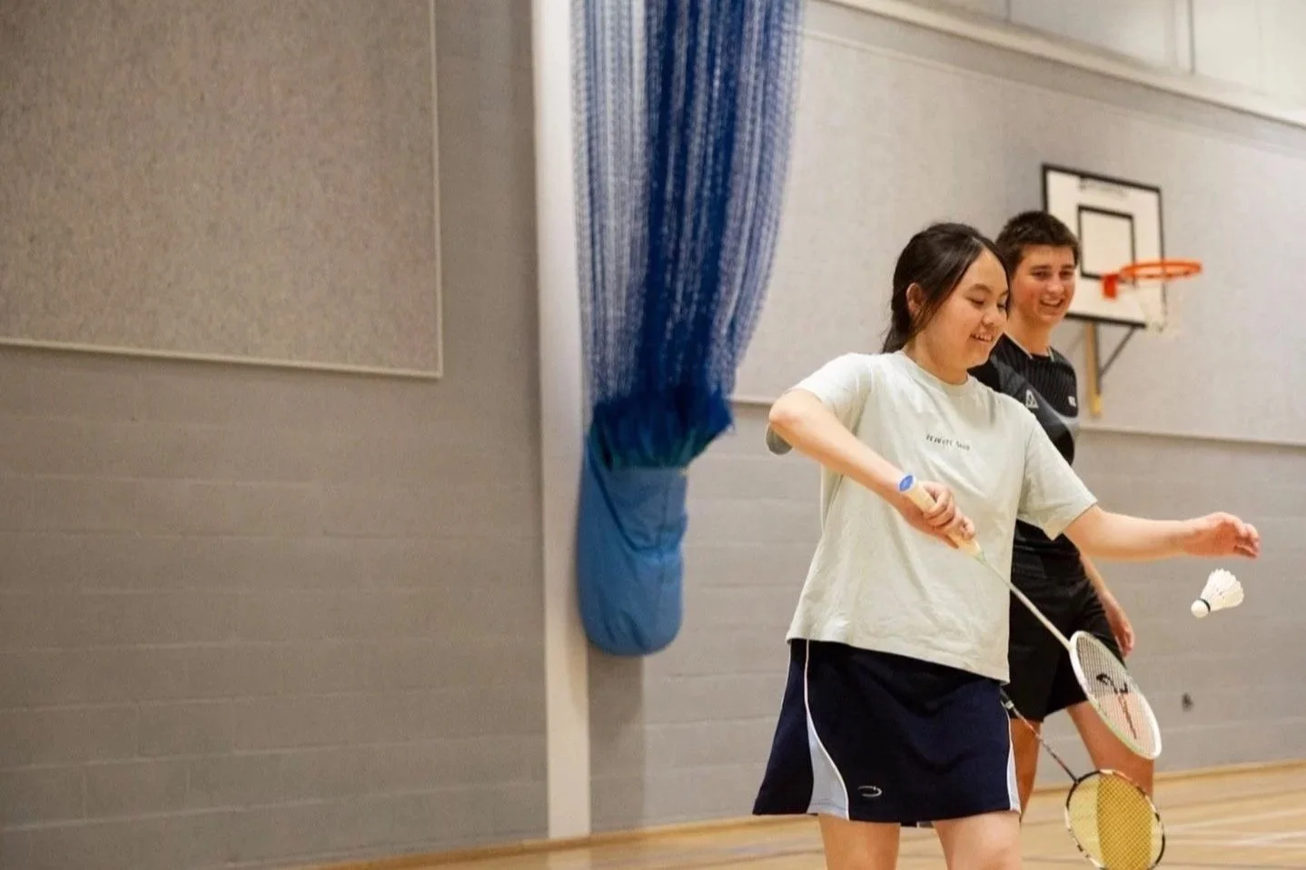 Two young women playing badminton indoors, one smiling and holding a racket, with a badminton shuttle in mid-air, in a gymnasium with a basketball hoop and blue curtain in the background.
