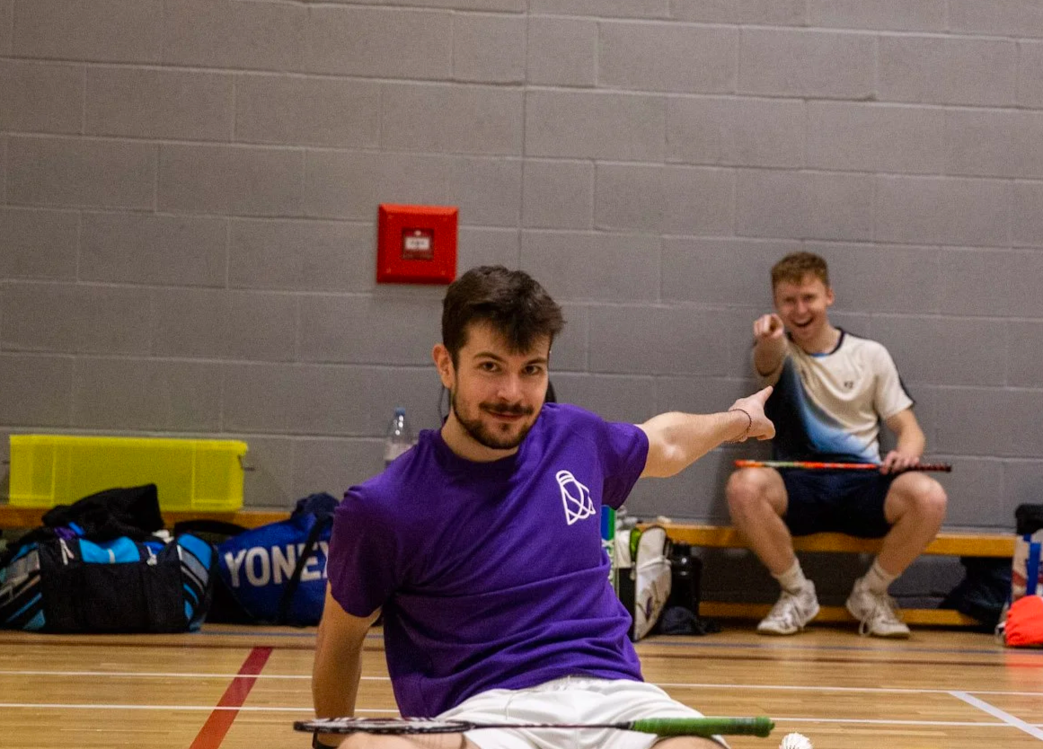 A young man with brown hair and beard smiling while standing against a beige cinder block wall, wearing a blue sports jersey with black and dark blue accents.