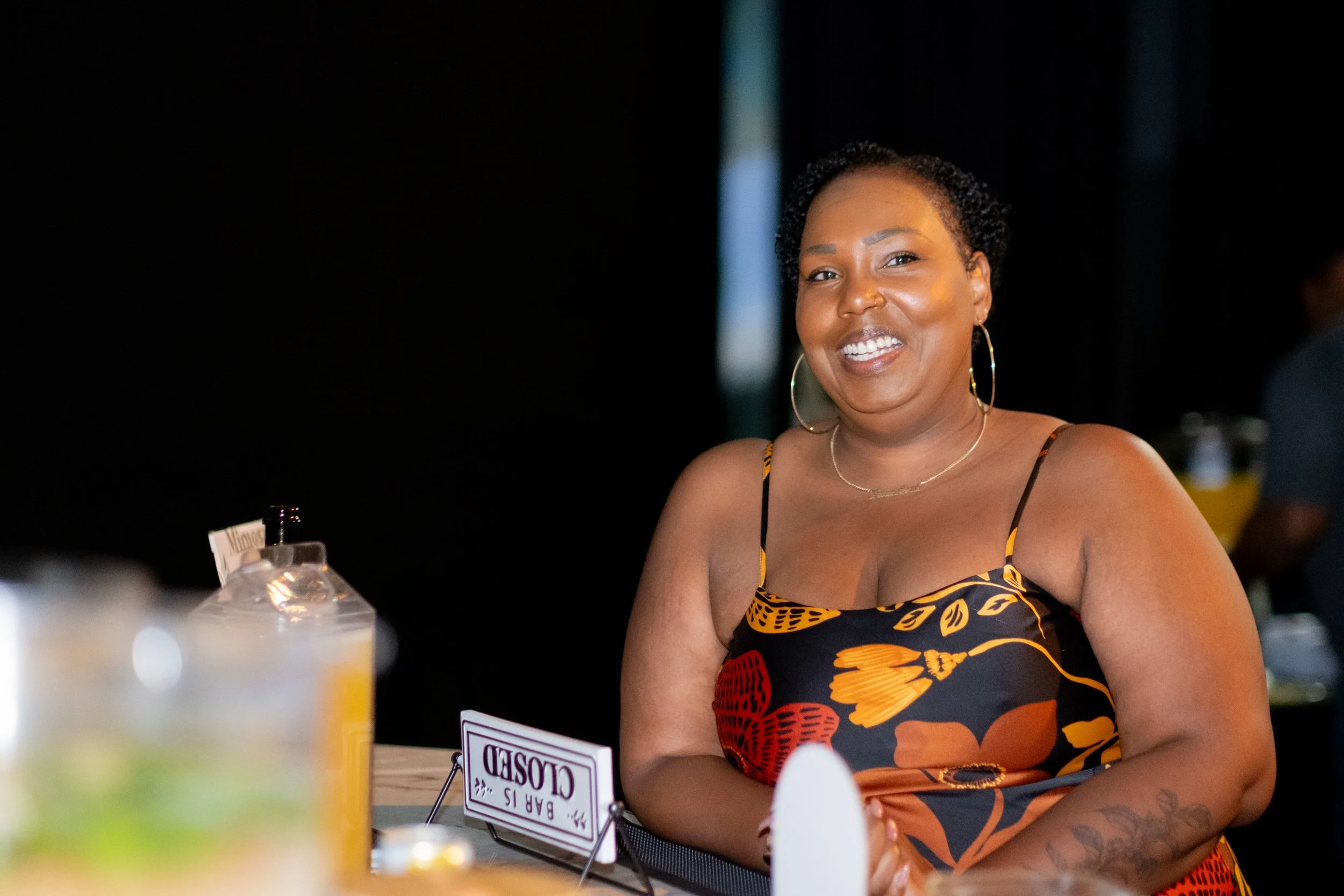 A woman smiles beside a table displaying a “BAR IS CLOSED” sign.