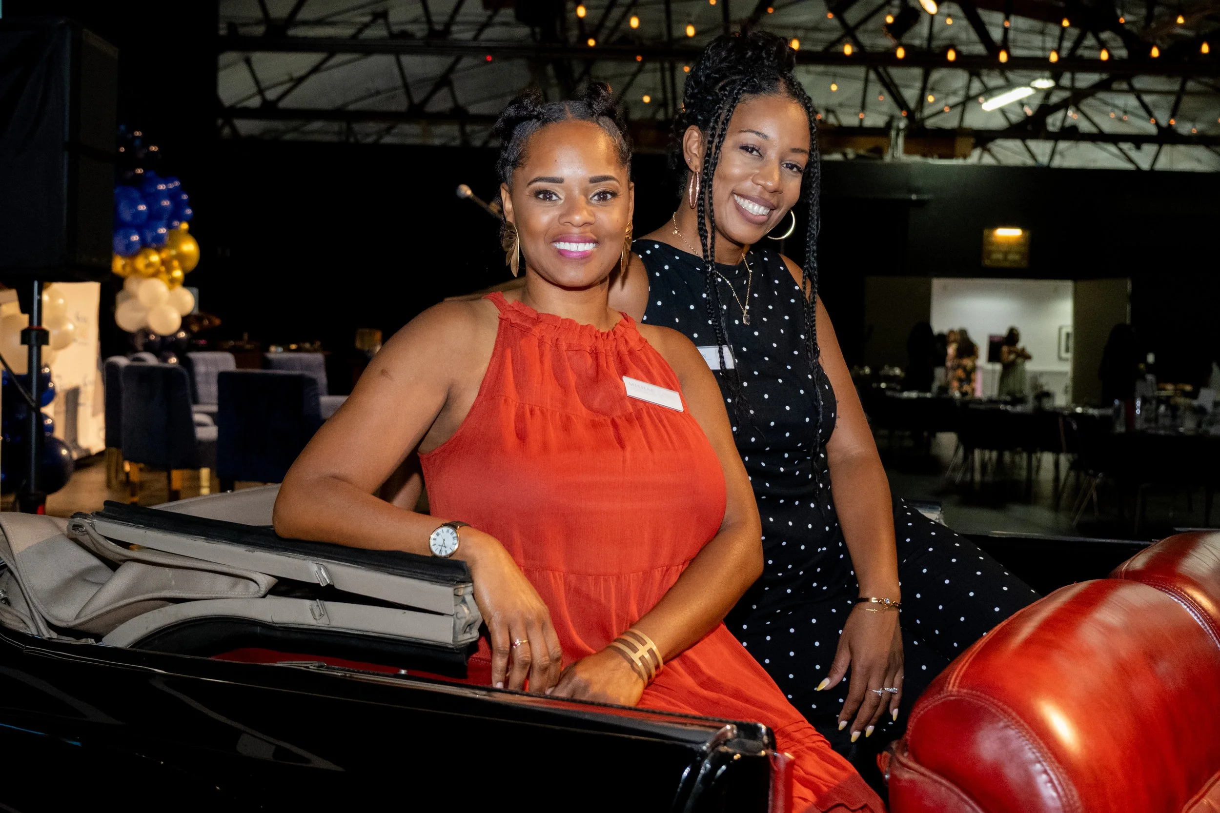 Two women smile while seated in a vintage black car at an indoor event.