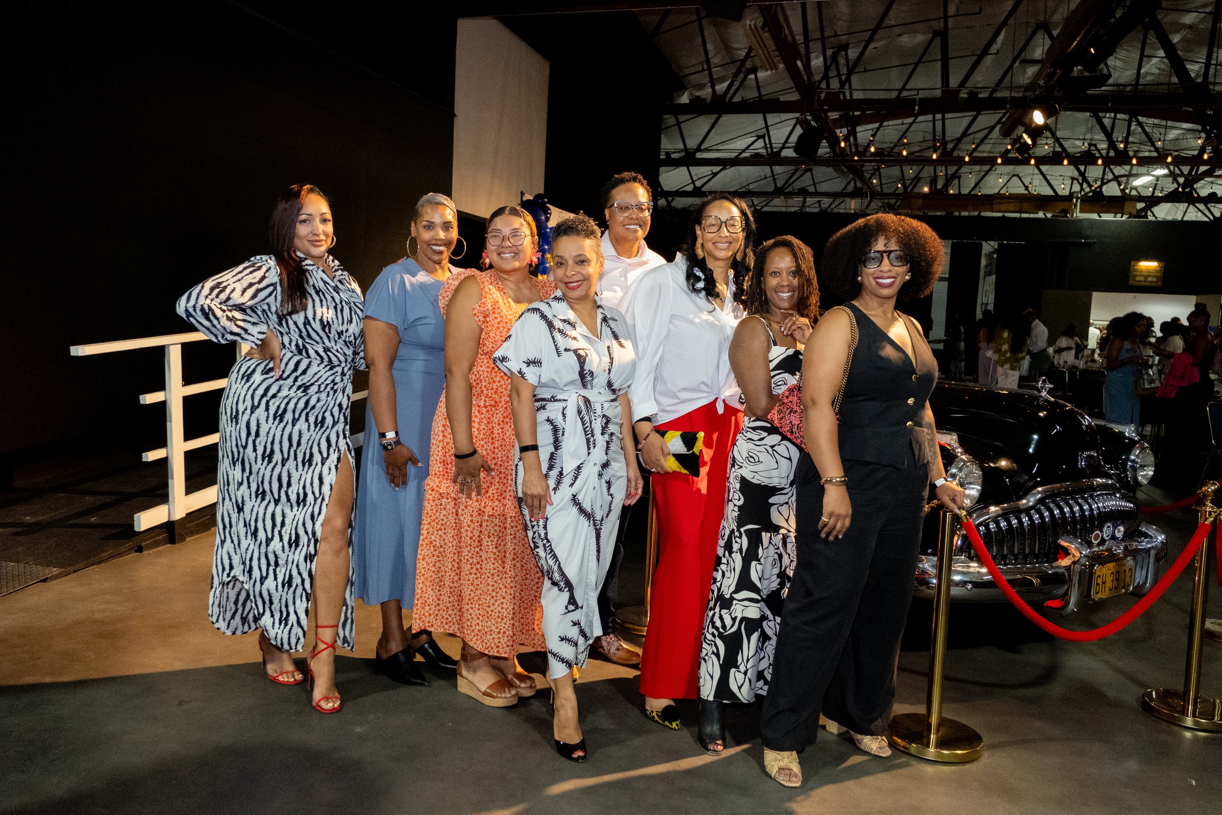 A group of eight women pose beside a vintage black car at an indoor event.