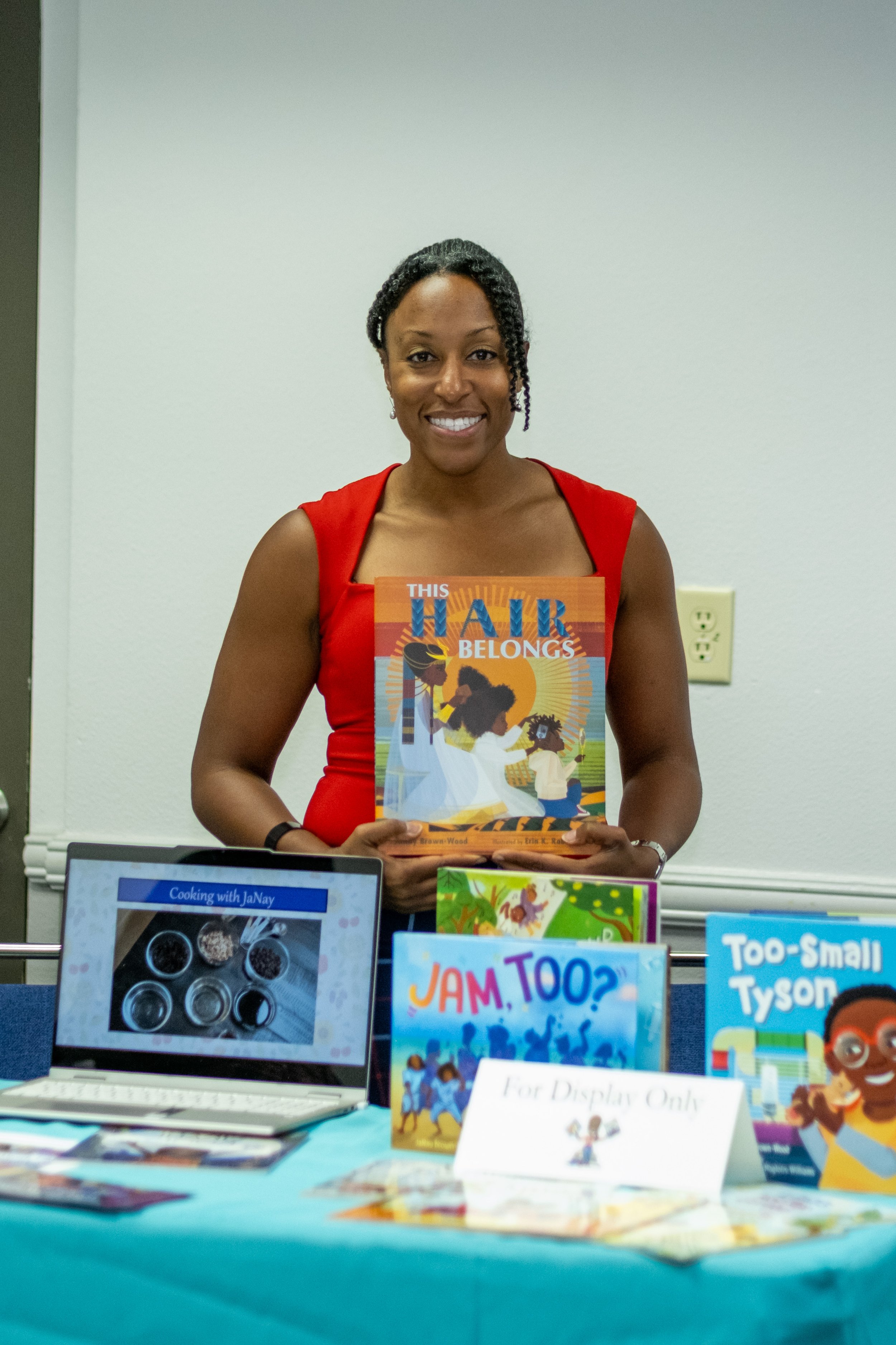 A woman holds a children’s book titled “This Hair Belongs” at a book display table.
