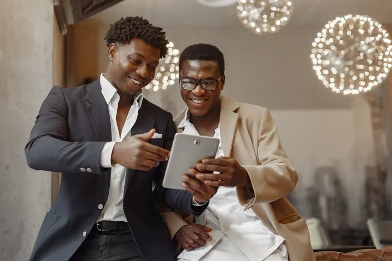 Two professionally dressed men smiling while looking at a tablet together in a modern office setting.
