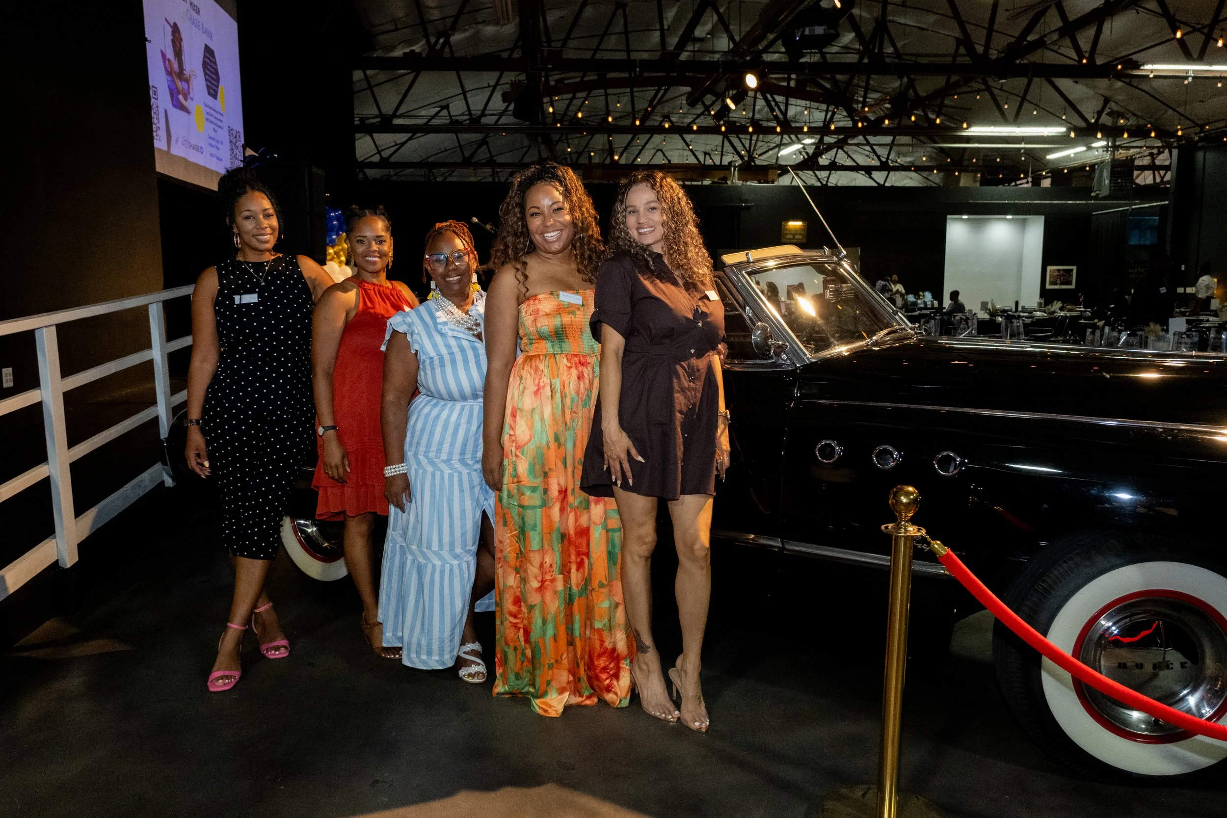 Five women stand beside a vintage black car at a branded brunch and mixer event.