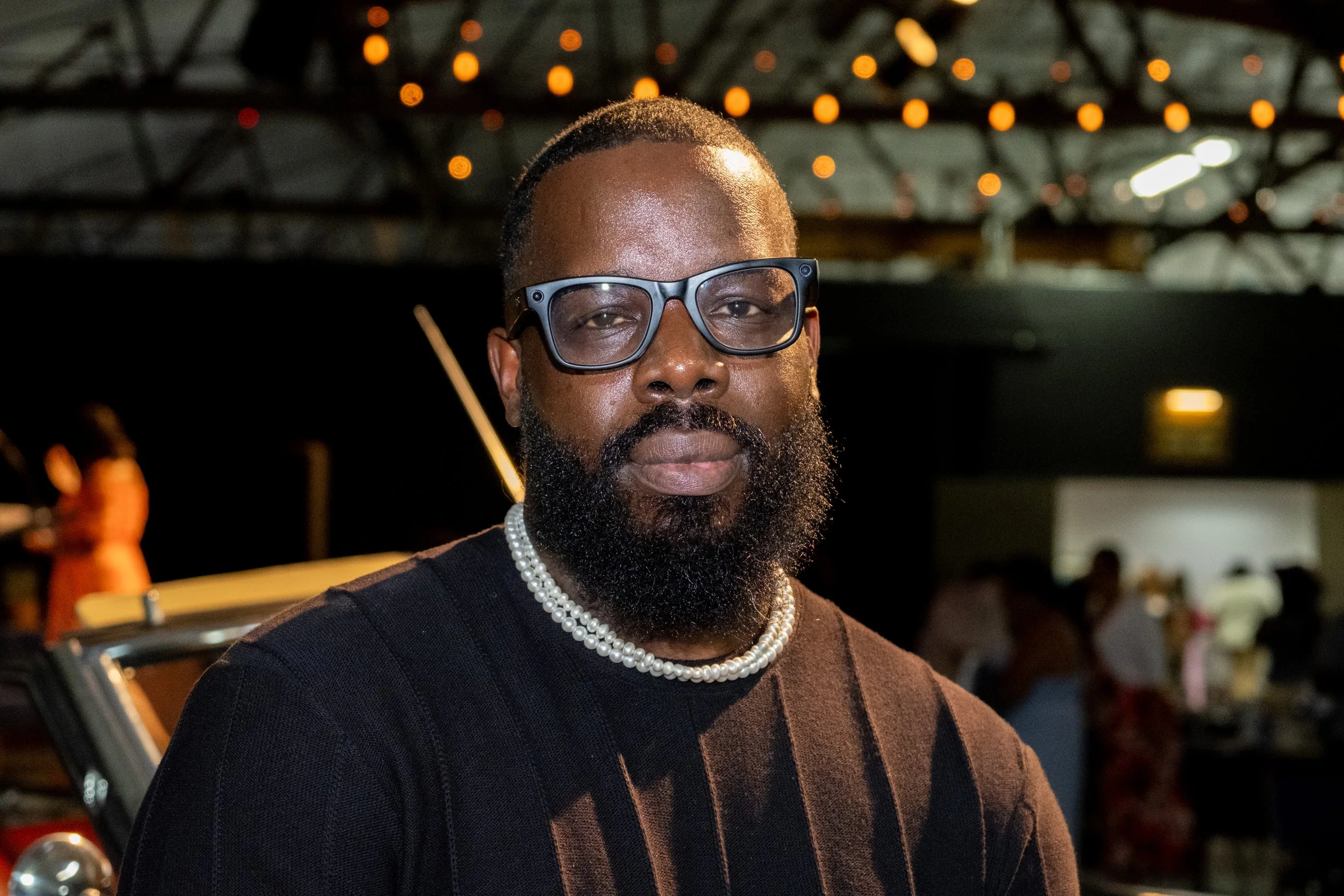 A bearded man wearing glasses and a pearl necklace smiles at the camera.