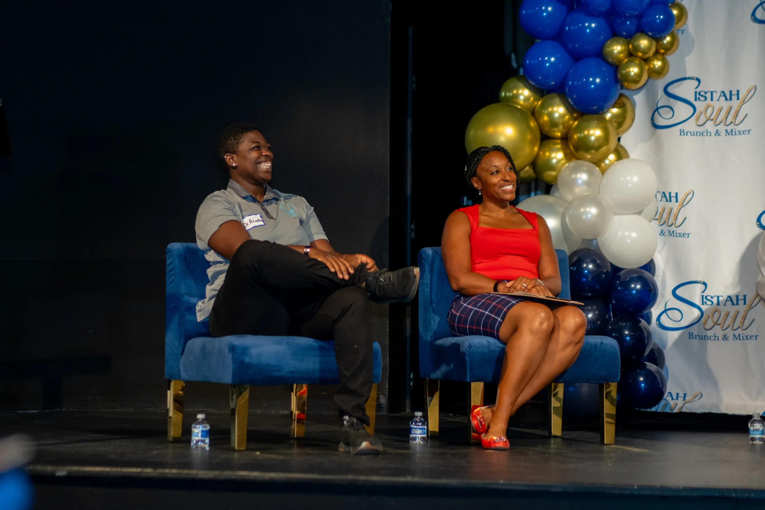 Two panelists sit in blue chairs and smile during a discussion.
