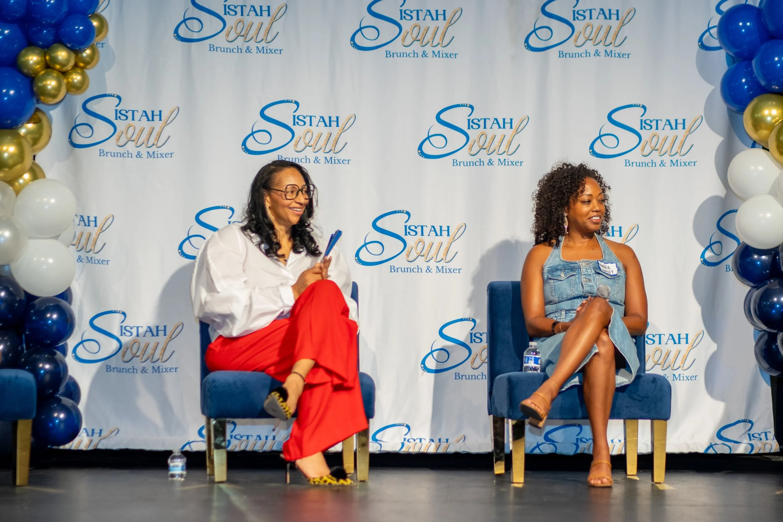 Two women sit on stage in front of a “Sistah Soul Brunch & Mixer” backdrop.