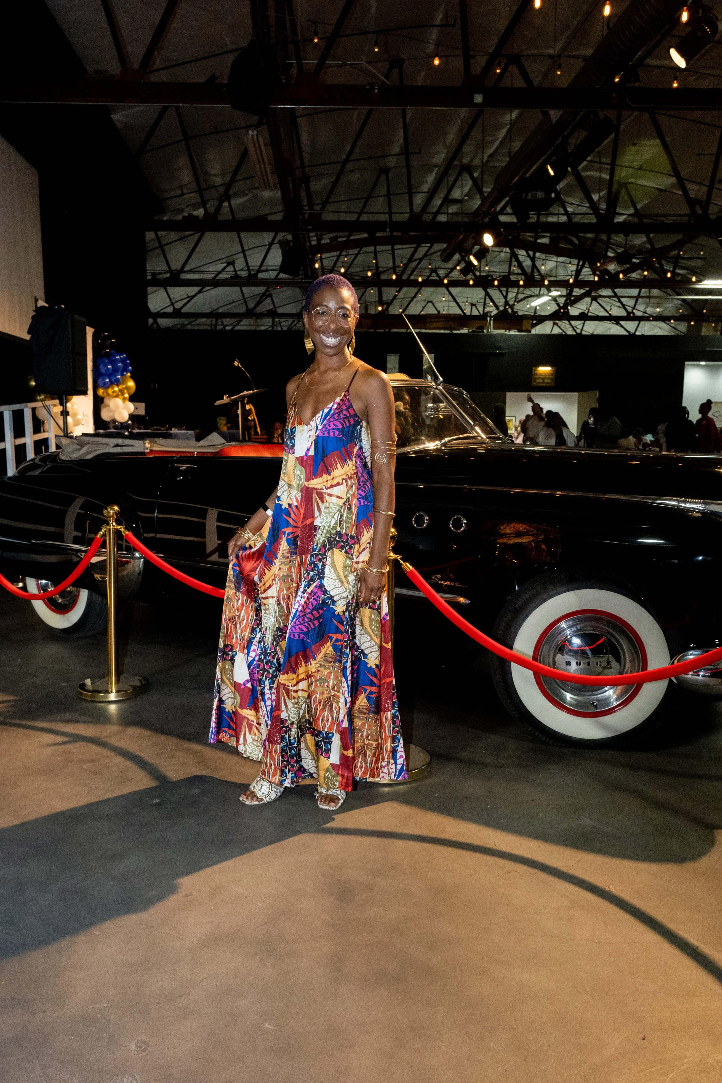 A woman in a colorful patterned dress poses beside a vintage black car.