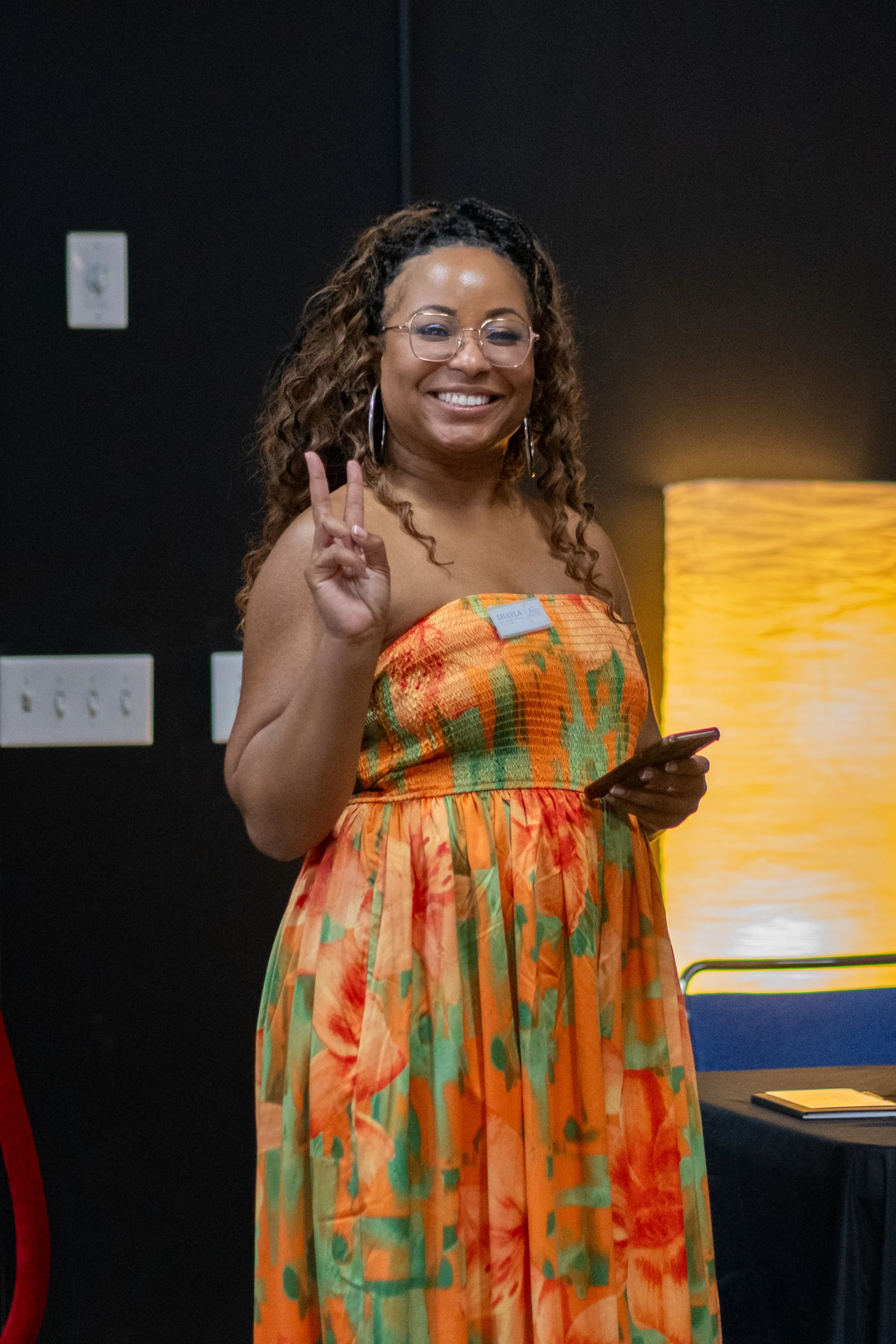 A woman in a colorful dress smiles and makes a peace sign while holding a phone.