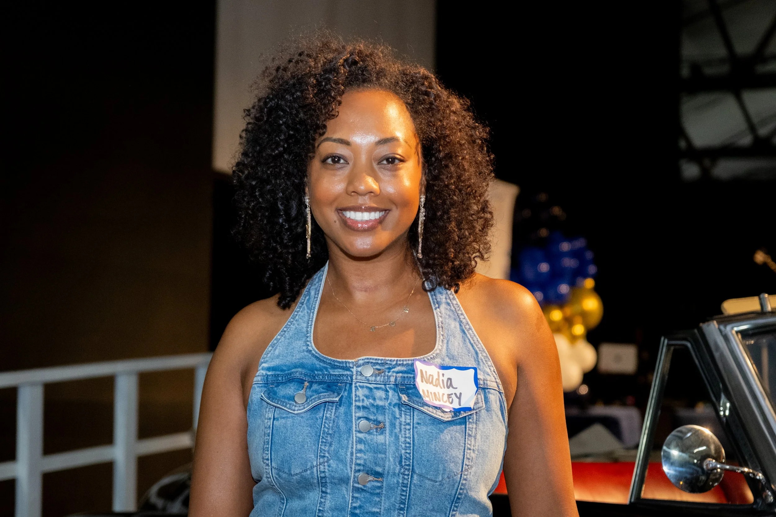 A smiling woman in denim overalls poses near a vintage car.