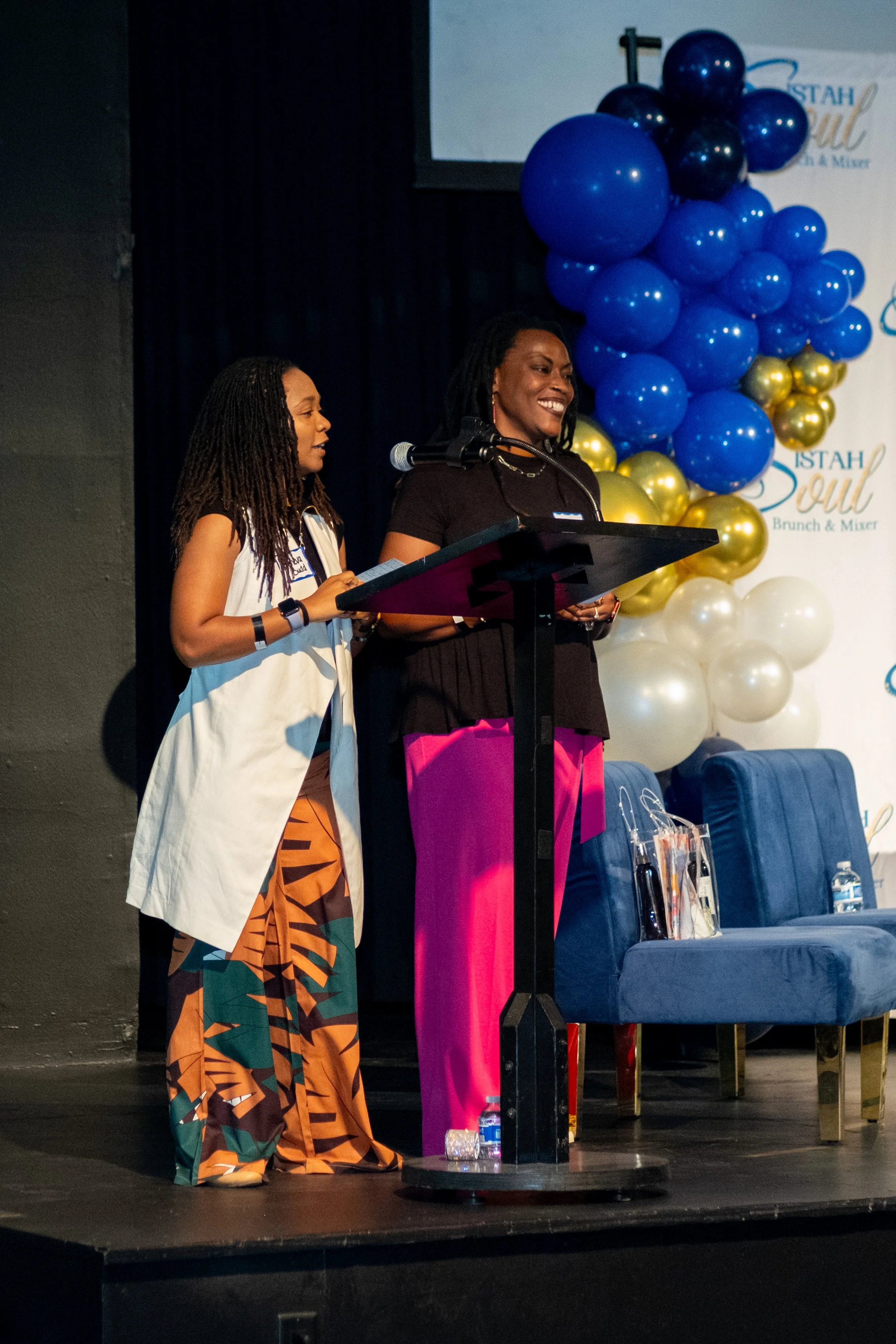 Two women stand at a podium in front of blue, gold, and white balloon decorations.