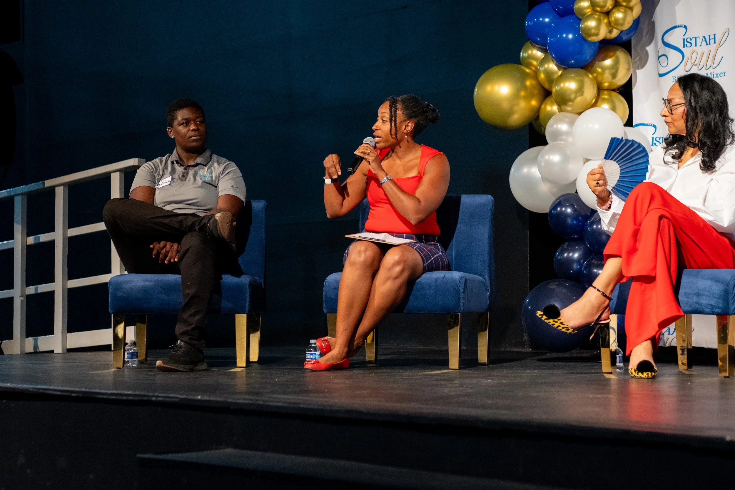 Three panelists sit on stage as a woman in a red dress speaks into a microphone.