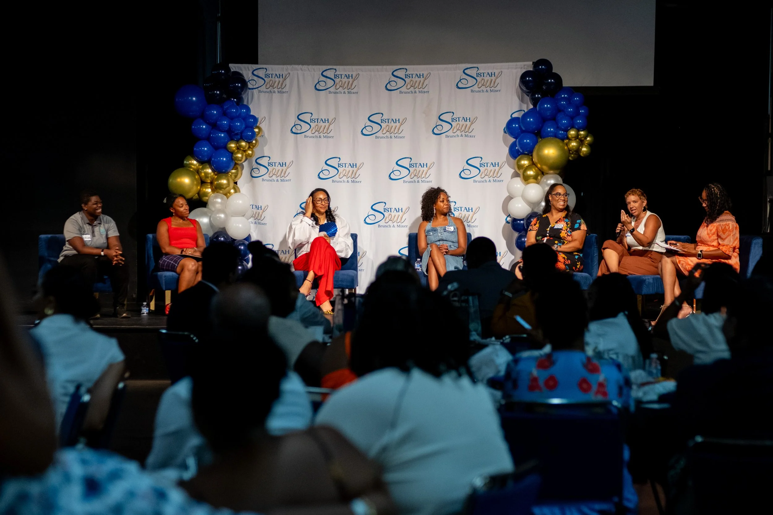 Seven panelists sit on stage in front of a “Sistah Soul Brunch & Mixer” backdrop while an audience watches.