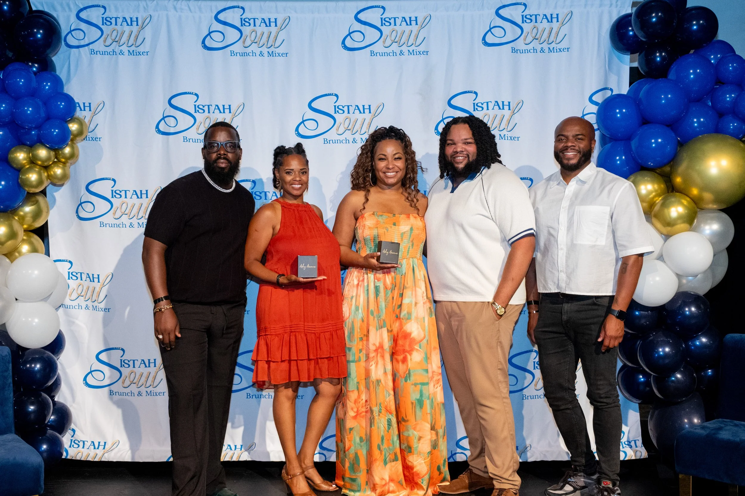 Five attendees pose in front of a branded backdrop; two women hold award boxes.