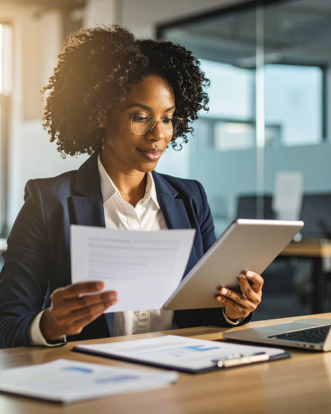 A women in a corporate setting at her desk holding a tablet in one hand and document in the other hand.